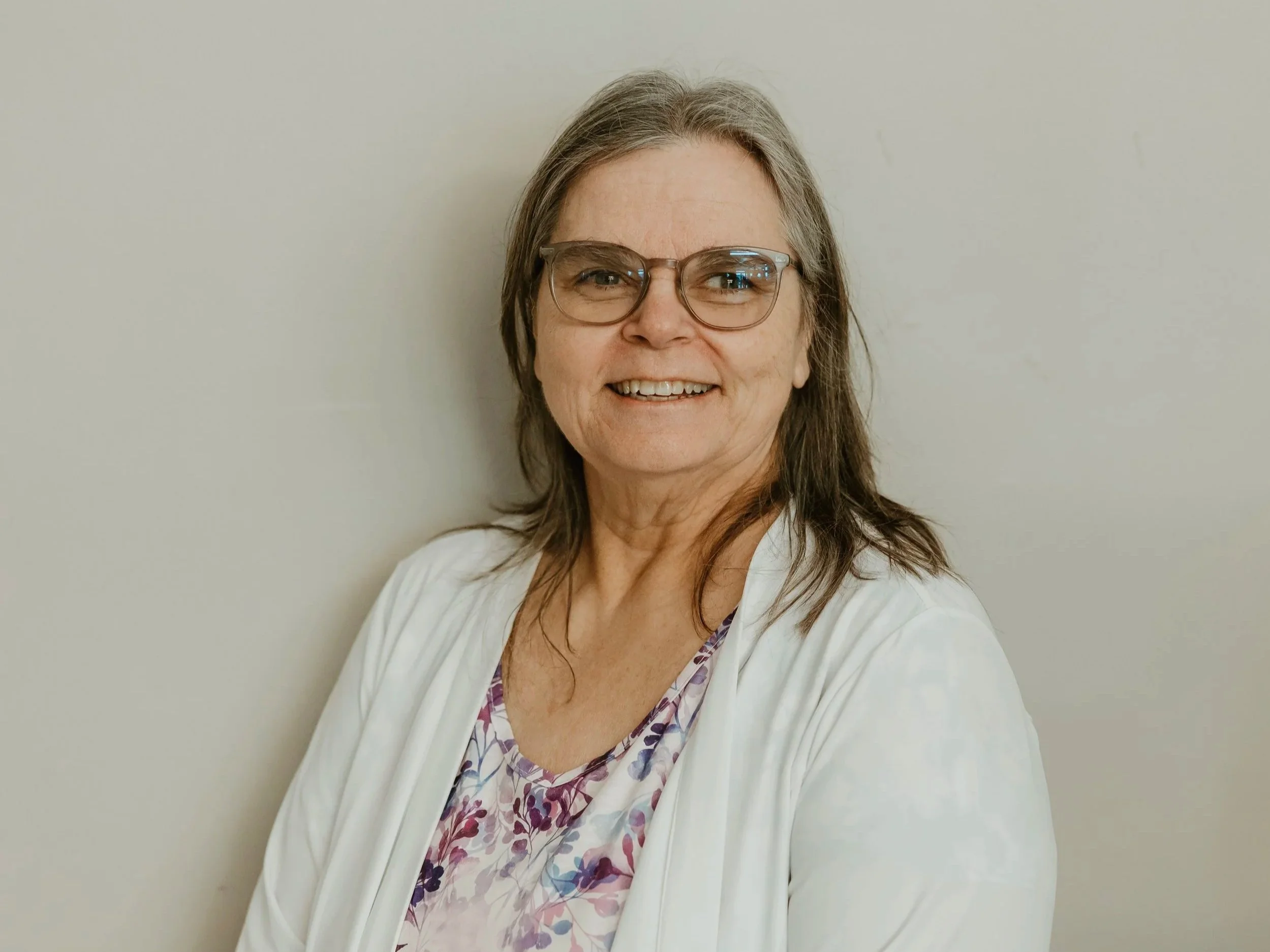 A middle-aged woman with long brown hair, glasses, and a warm smile, standing against a plain light-colored wall, wearing a white coat over a floral-patterned top.