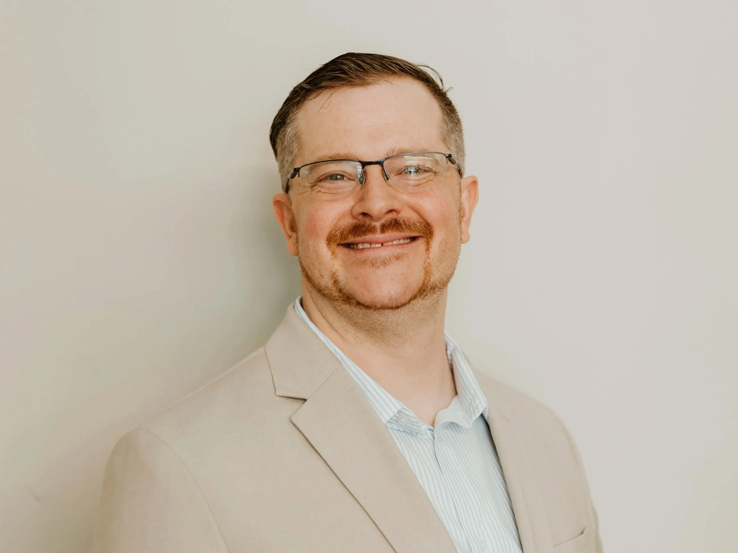 A smiling man with glasses, light brown hair, and a beard wearing a beige suit jacket and a light striped shirt, standing against a plain off-white background.