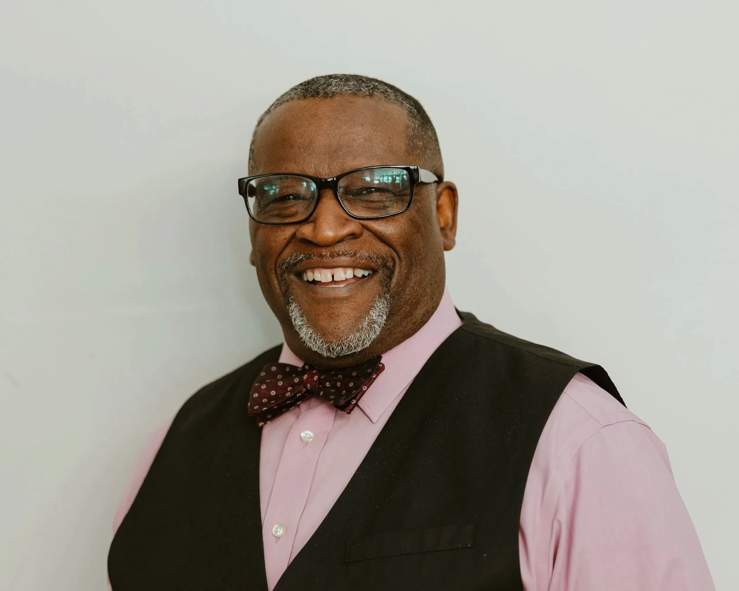 Smiling middle-aged man with short gray hair, wearing black glasses, a pink dress shirt, a dark vest, and a maroon bowtie with small white polka dots, against a plain light background.