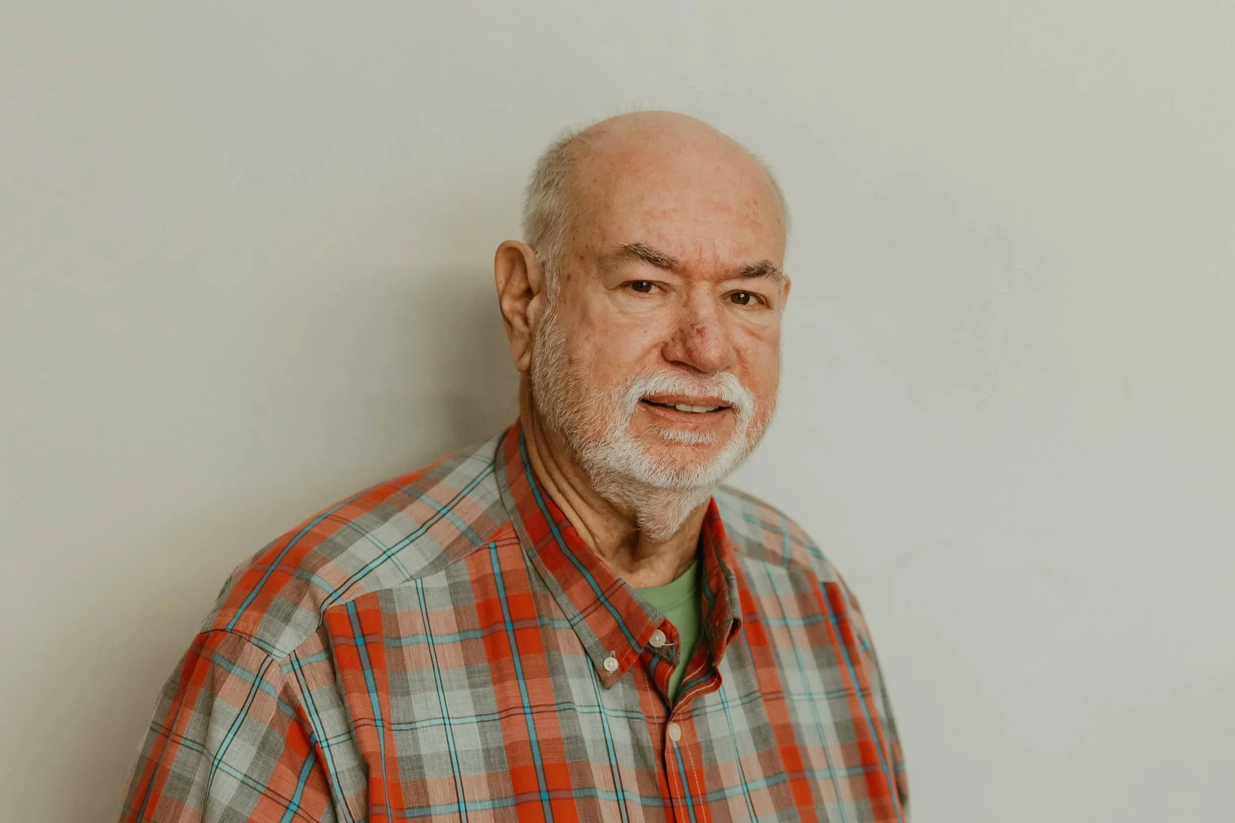 An older man with a beard and mustache, wearing a red and gray plaid shirt, standing against a plain light-colored wall.