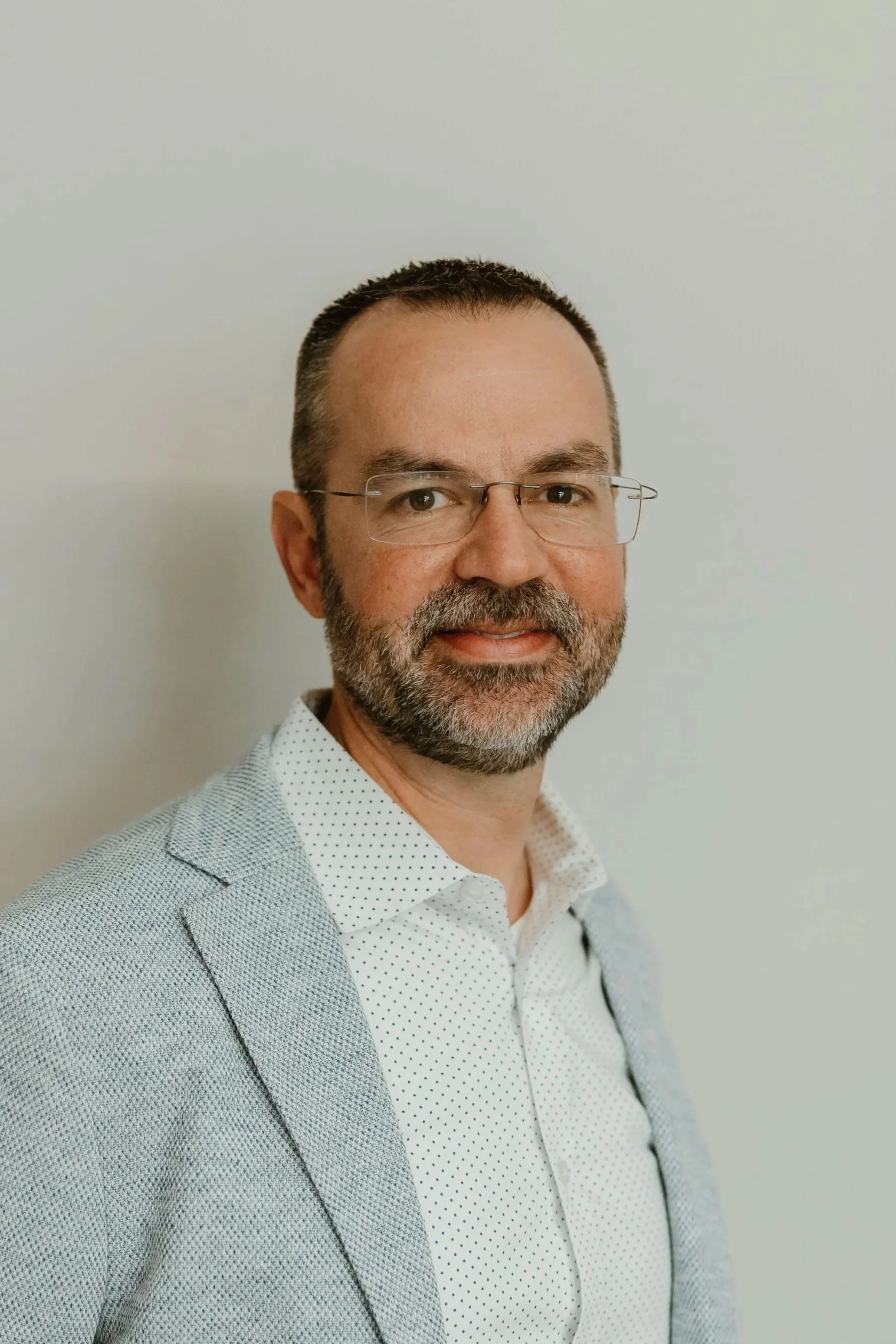 A middle-aged man with short dark hair, a beard, and glasses, wearing a light grey blazer and a white dress shirt with small black dots, smiling, standing against a plain, light-colored wall.