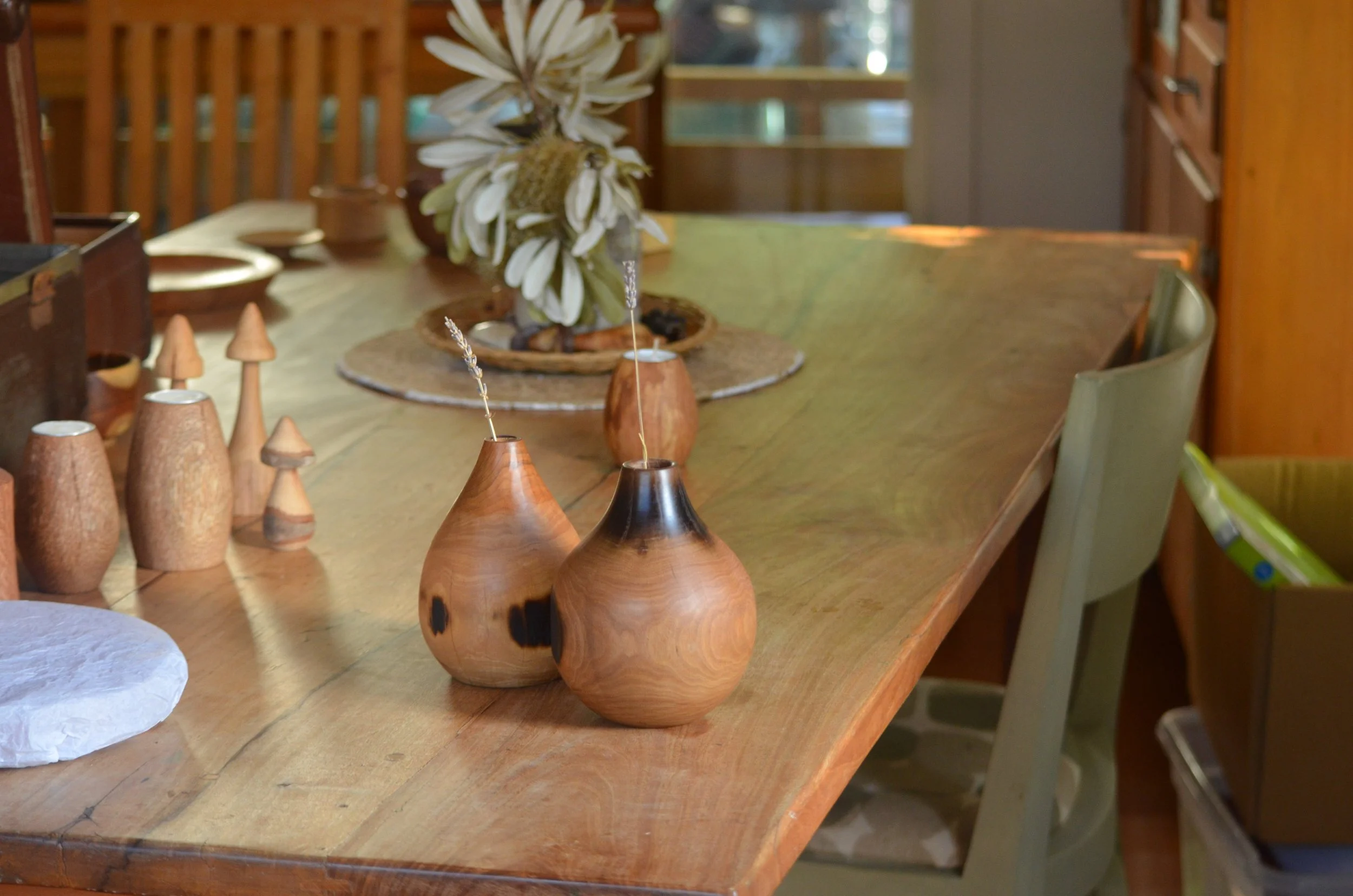 A wooden dining table decorated with minimalist wooden vases, some with small dried flowers, and a centerpiece with a large white flower arrangement. The background shows a buffet with more wooden decor and a window letting in natural light.