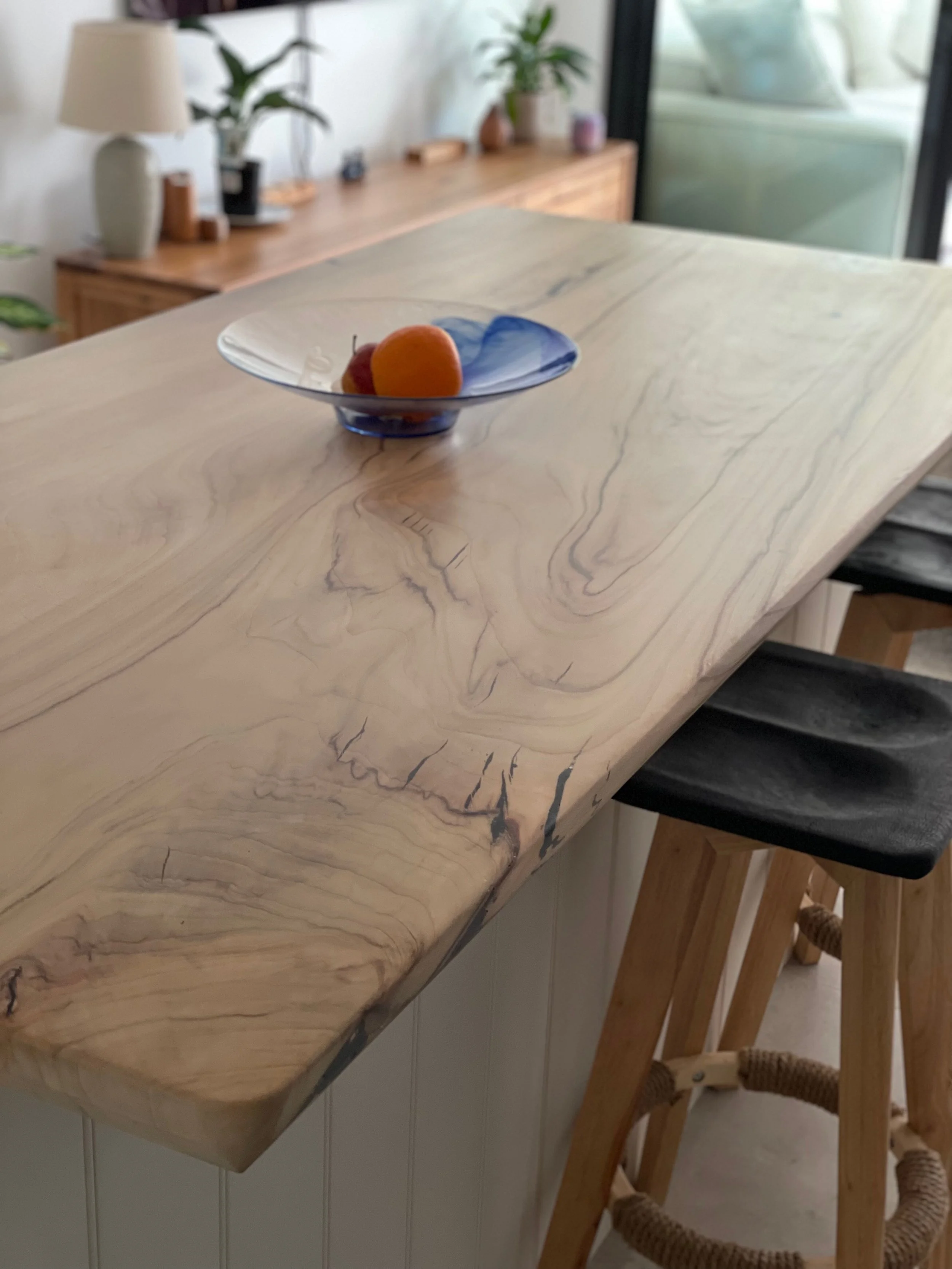 Close-up of a light-colored wooden table with a blue glass bowl containing two oranges and a red apple, in a room with houseplants, a wooden sideboard, and a sliding glass door.