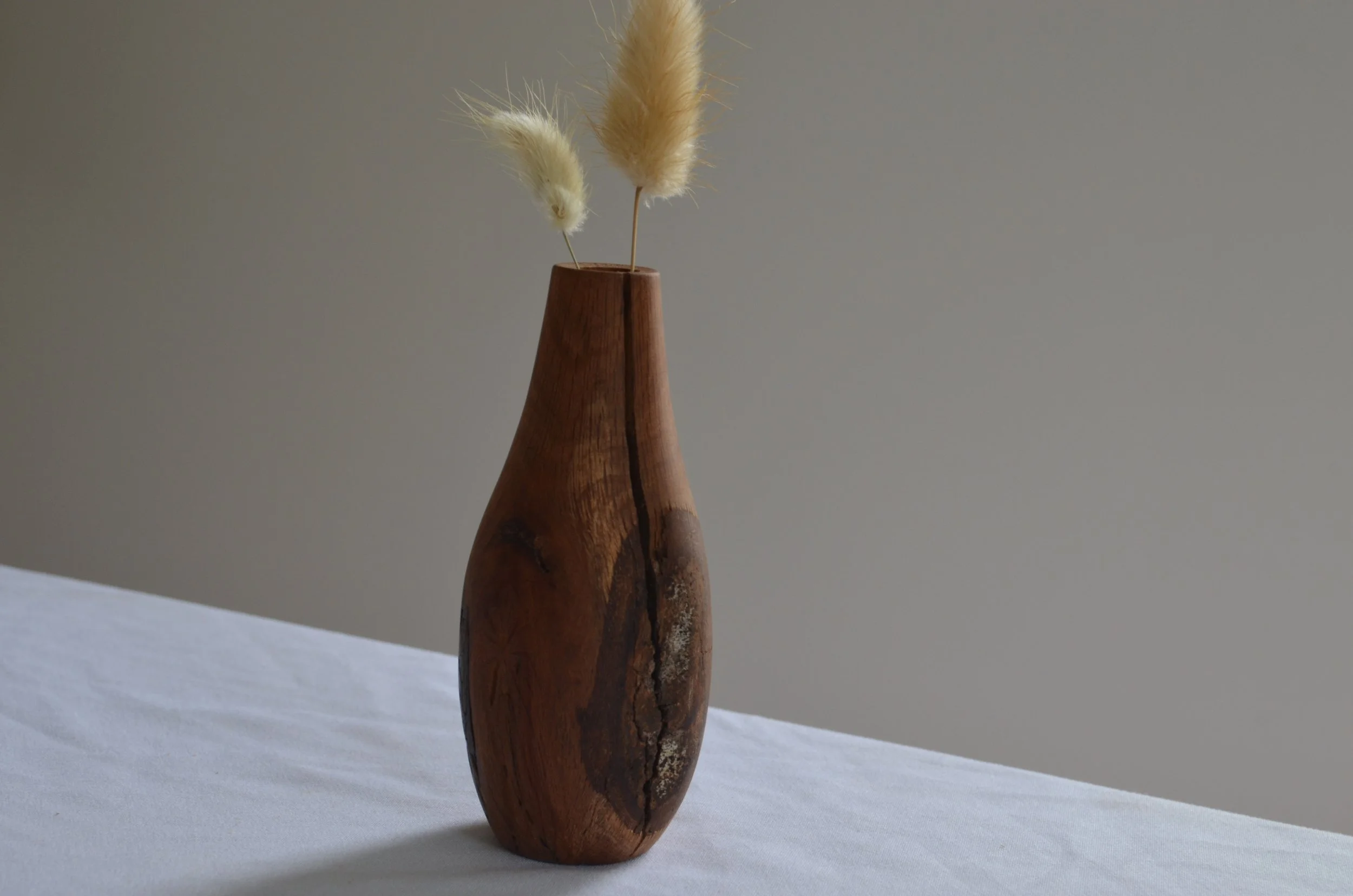 Wooden vase with two dried pampas grass stems on a white table against a plain gray background.