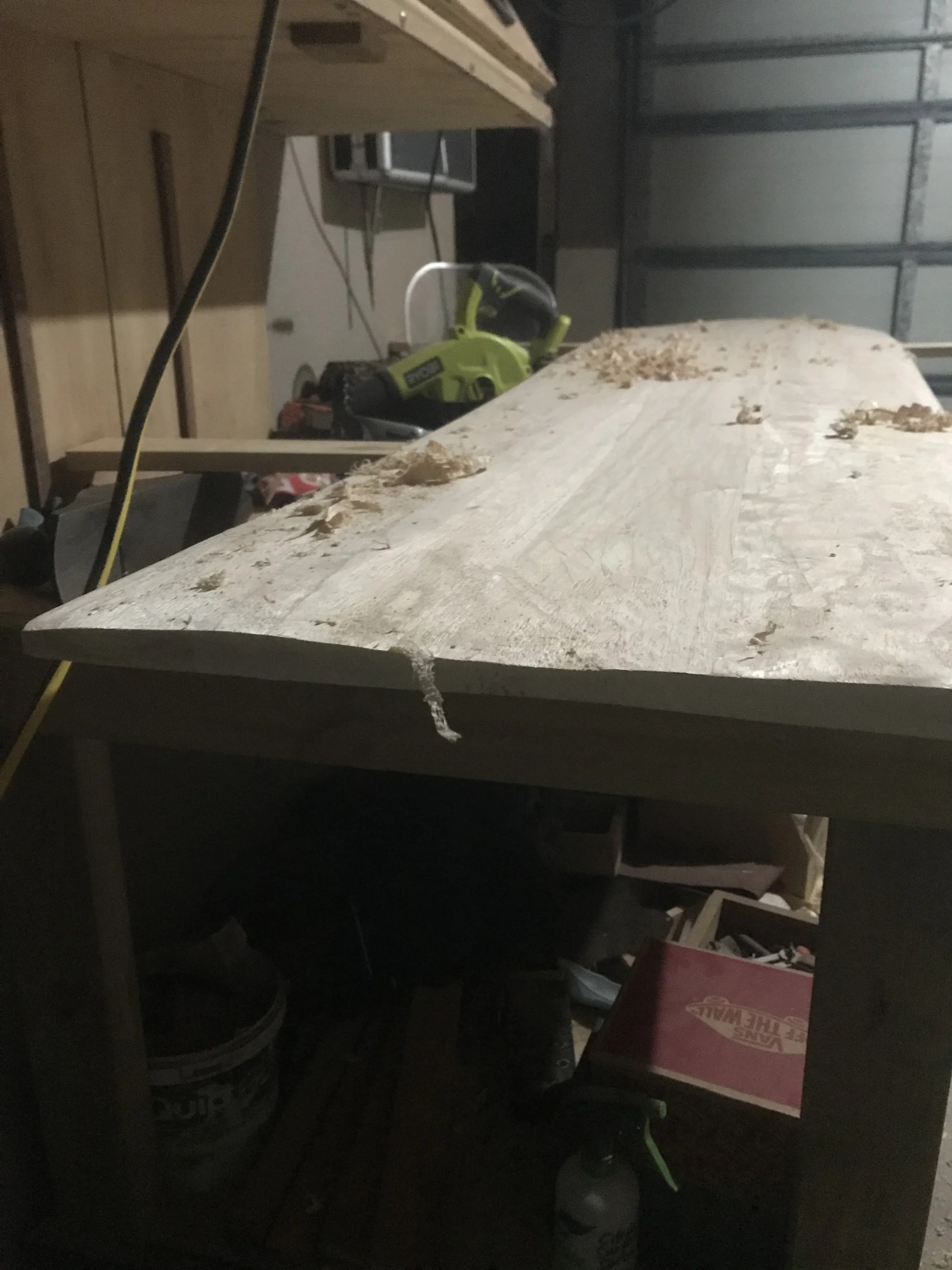 Woodworking shop with a large piece of wood on a workbench, power tools, and supplies in the background.