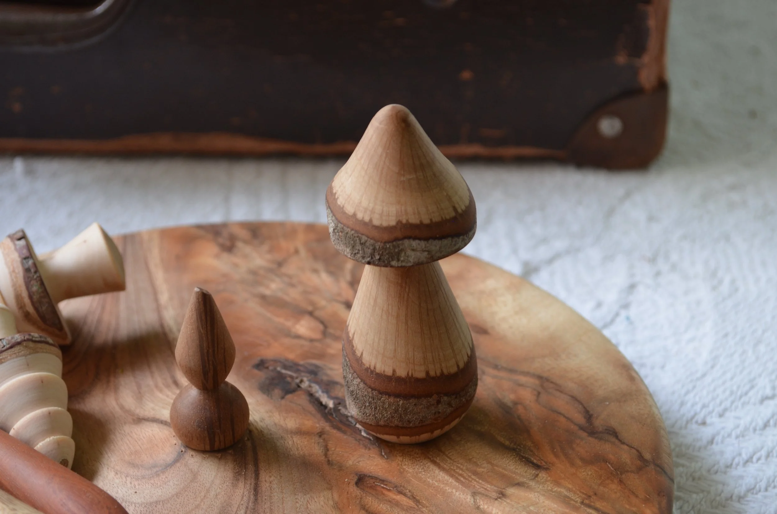 Wooden mushroom and smaller wooden sculpture on a round wooden surface, with a textured white fabric and a dark, partially visible object in the background.