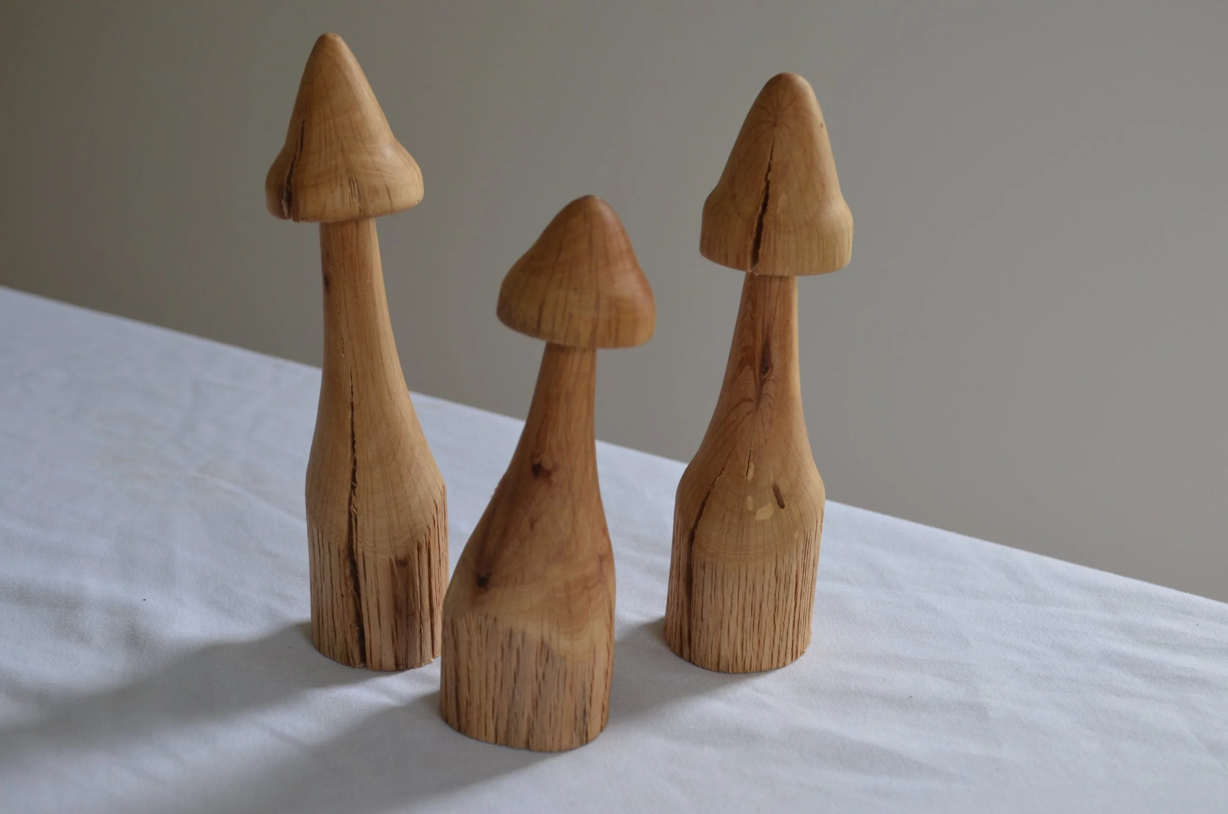 Three wooden mushroom-shaped sculptures on a white cloth surface with a plain gray background.