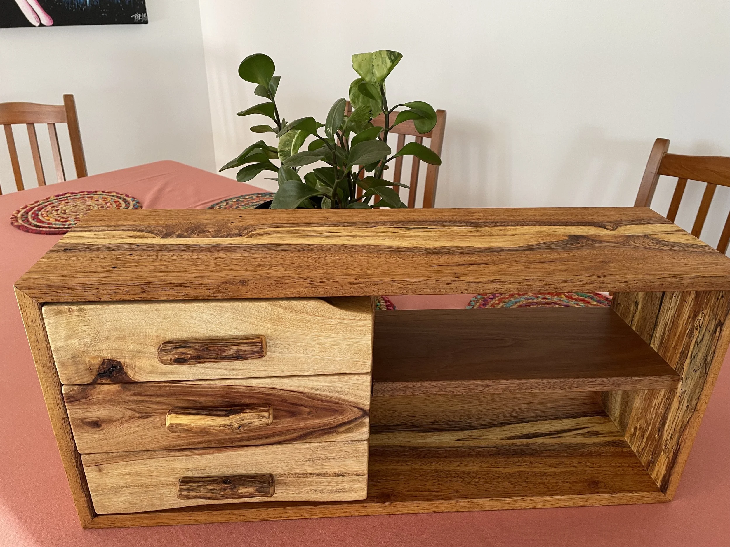 A wooden desk with three small drawers on the left side and two open shelves on the right side, placed on a pink tablecloth in a dining area with wooden chairs and a potted plant in the background.