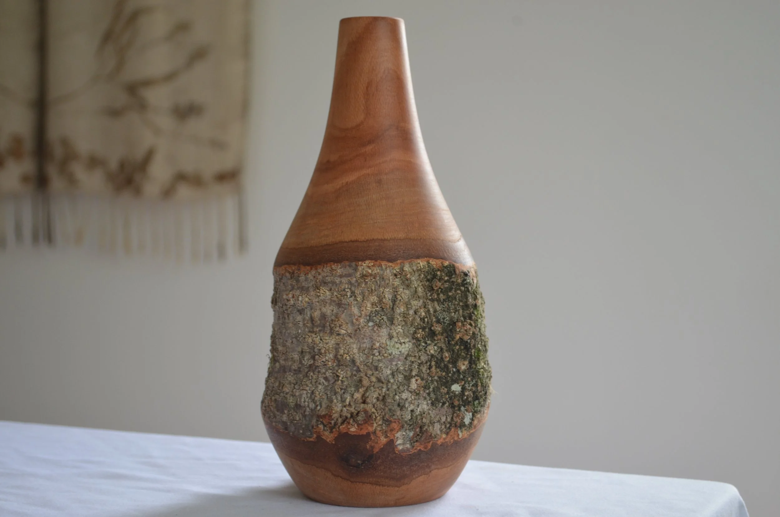 Wooden vase with a rough bark-covered middle section placed on a white tablecloth, against a neutral background.