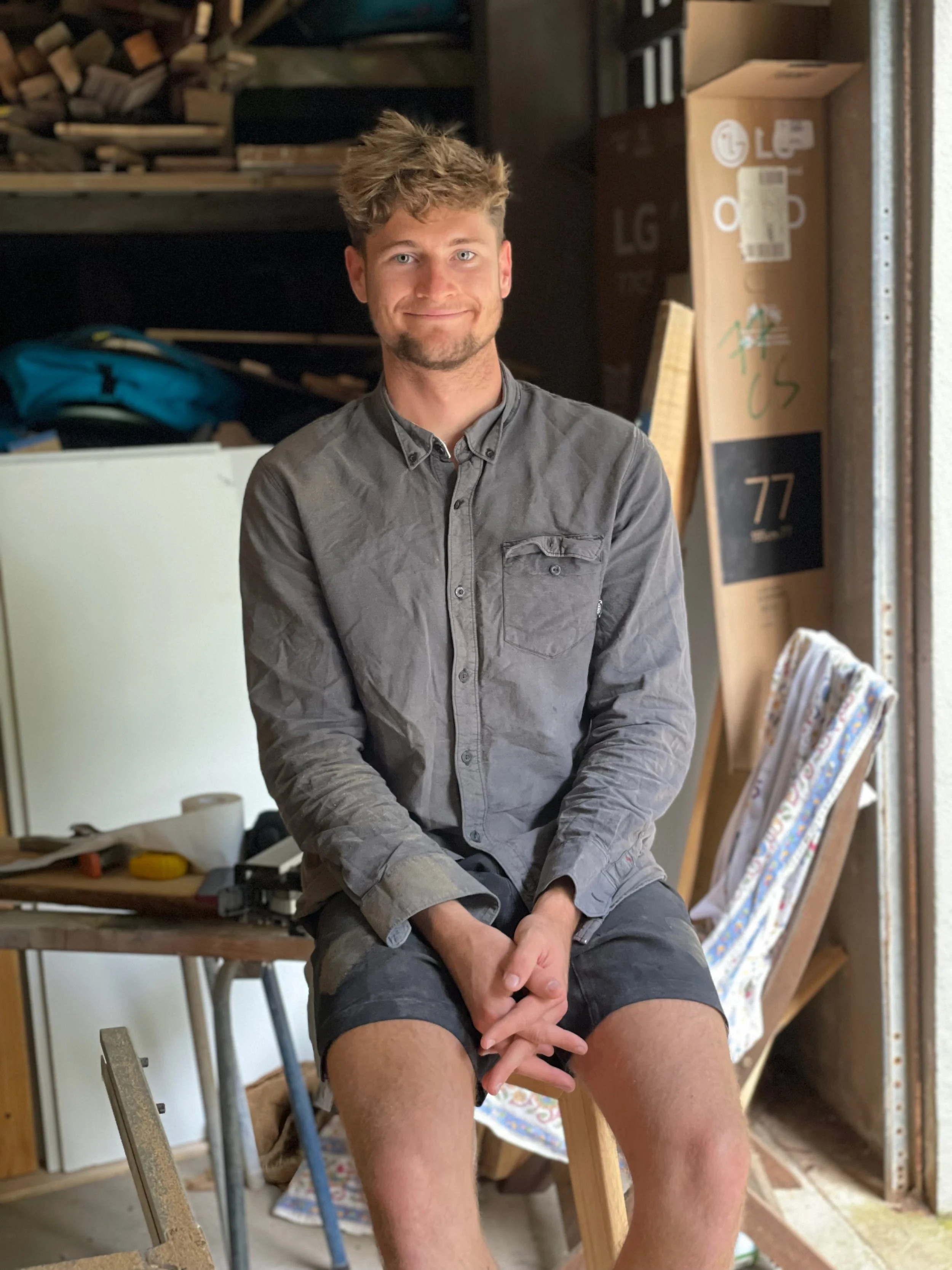 A young man with light skin, tousled hair, and a slight beard, sitting on a stool in a cluttered workshop or storage area, smiling at the camera, wearing a gray button-up shirt and dark shorts.