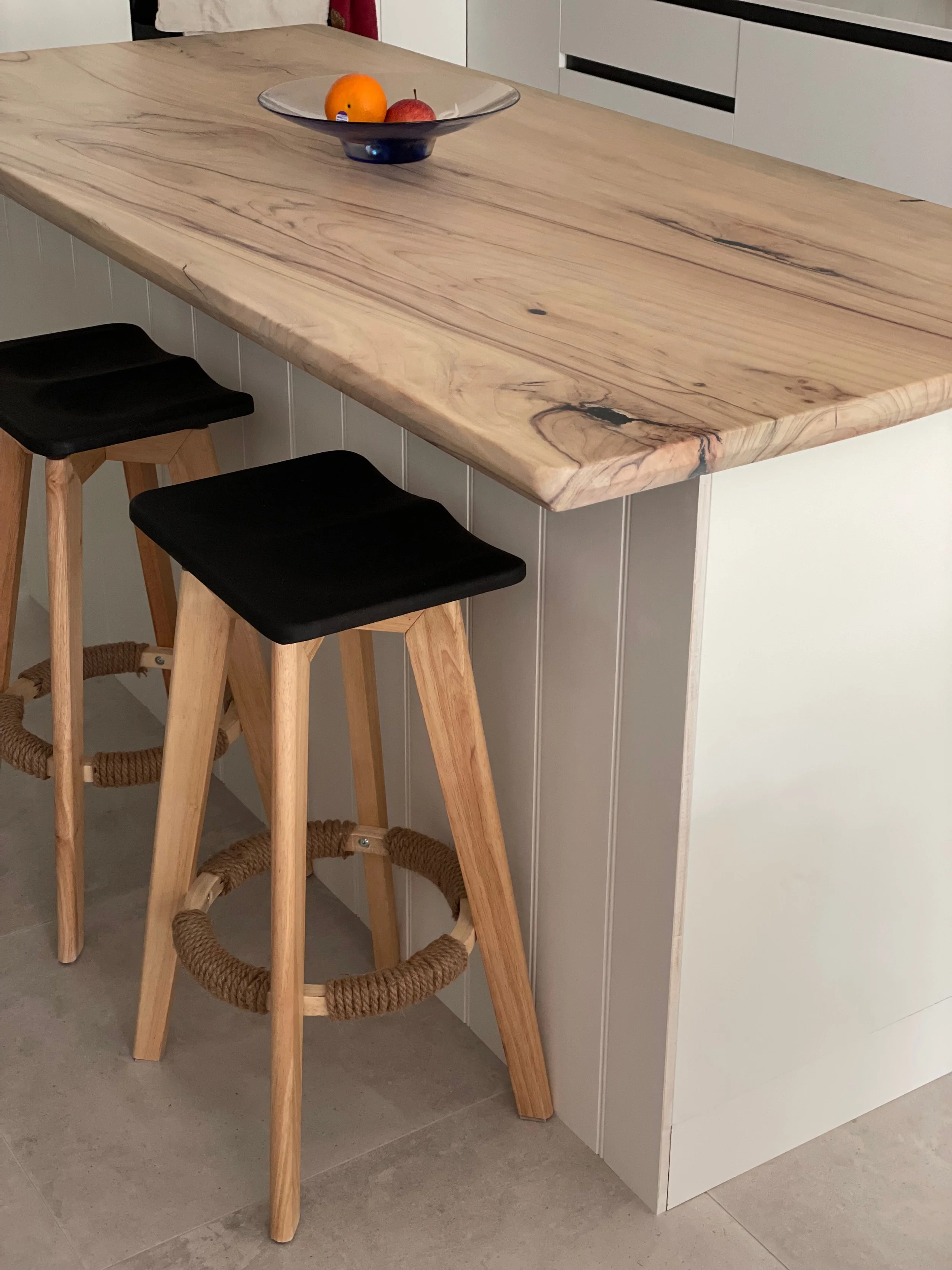 A kitchen island with a light wood countertop and two black and wood bar stools with rope footrests. On the countertop is a glass bowl containing oranges and apples.