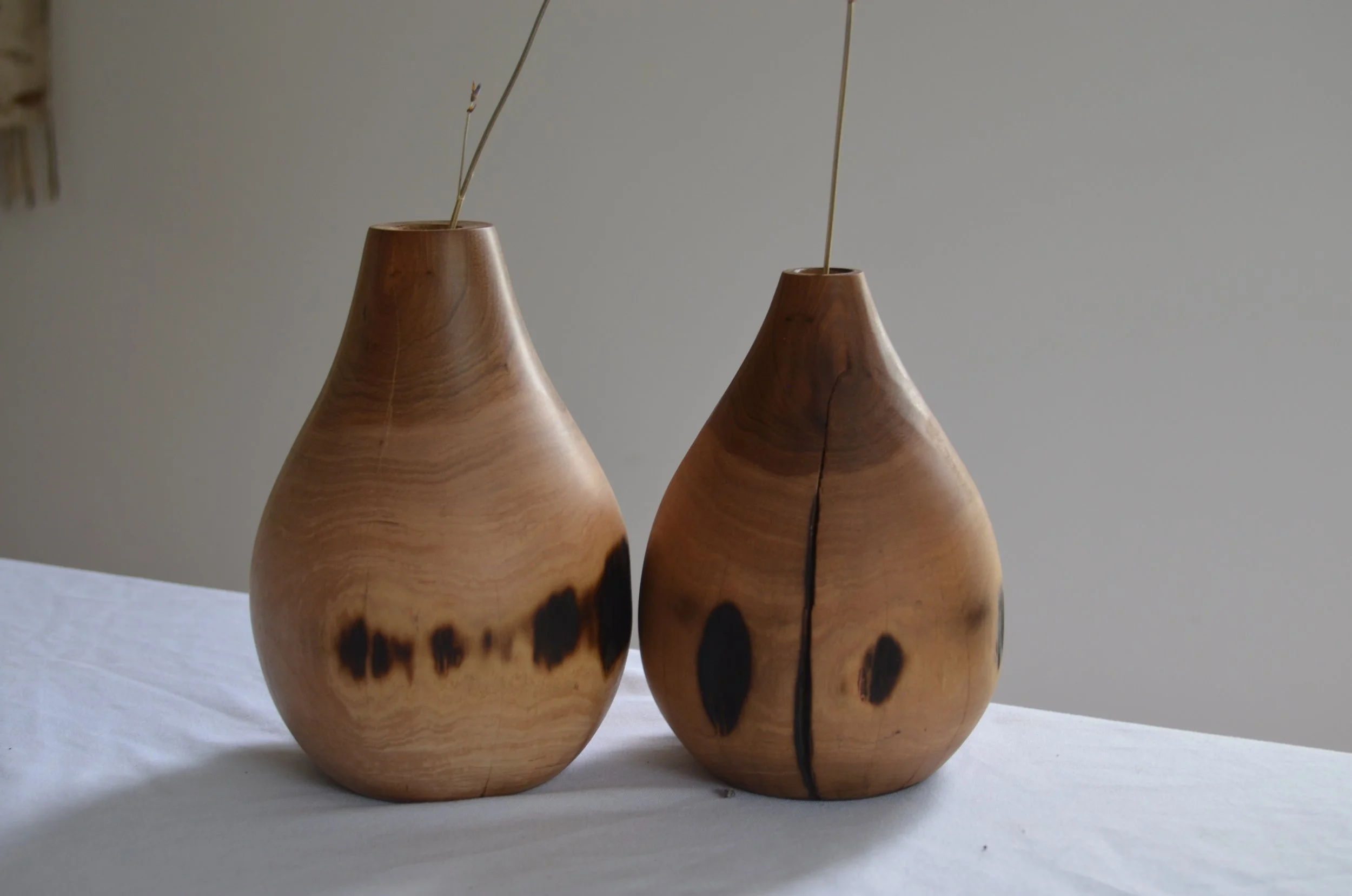 Two wooden vases with natural wood grain and dark spots, placed on a white surface against a plain light-colored wall.