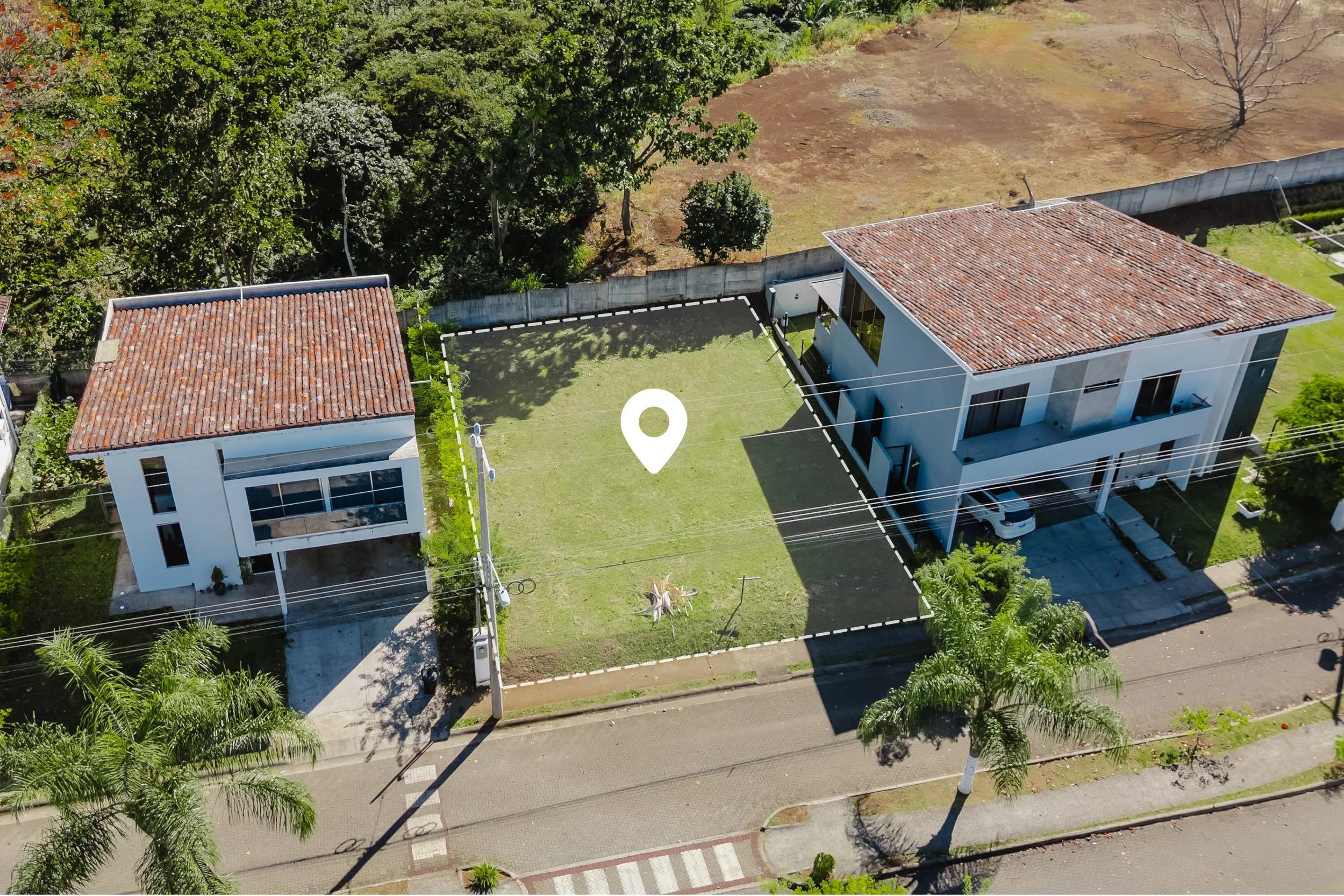 Vista aérea de dos casas lado a lado en un vecindario, con un lote de césped vacío en medio, arboles y una calle frente a ellas.