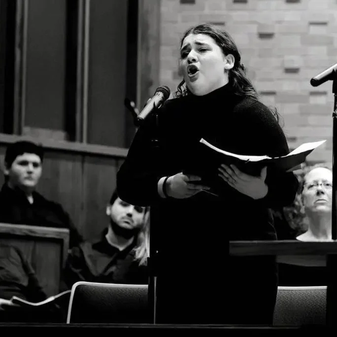 Black and White Photo of Woman singing into a microphone. She is wearing a black turtle neck and is holding sheet music. Her face and the faces of the people sitting behind her look somber