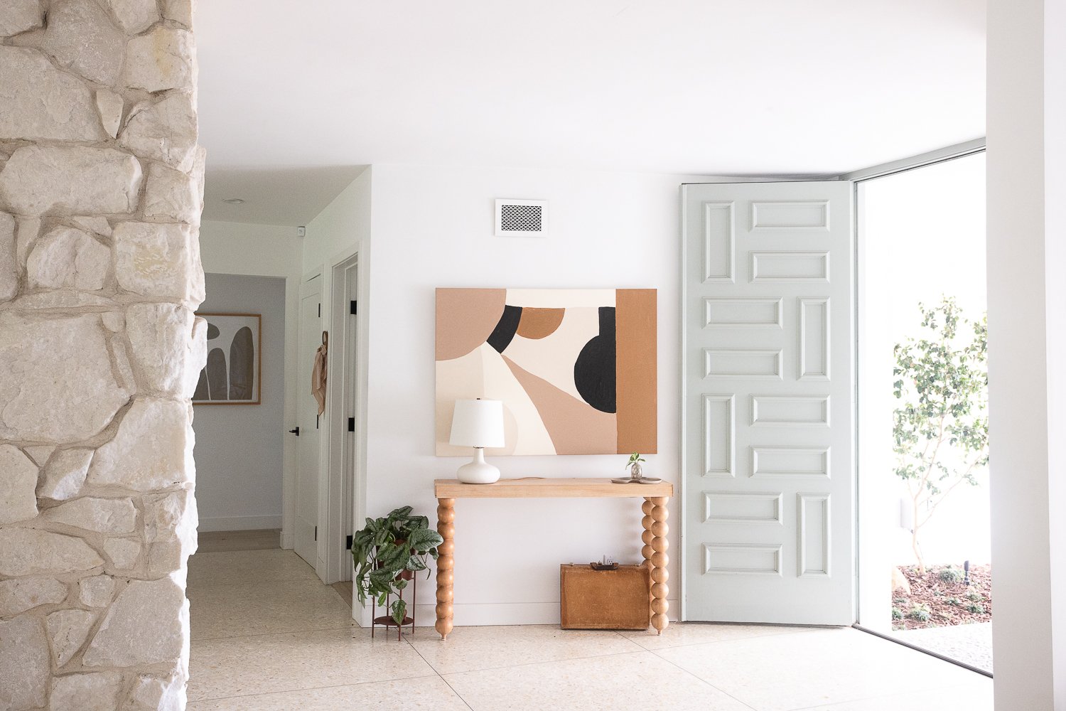 An entryway with a beige stone wall on the left, a white wall in the center with an abstract art piece, a light wood console table with round legs, a white table lamp, a small plant, a brown suitcase underneath, and an open door with a window to the 