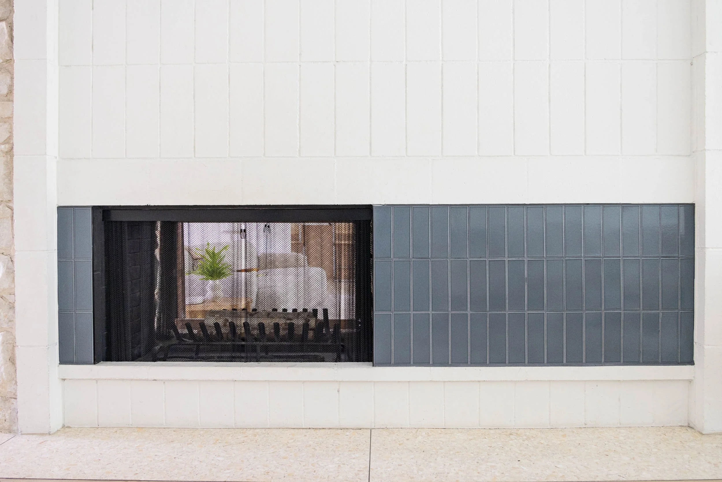 Fireplace with a black metal grate, surrounded by white and gray tiles, with a potted plant visible through the glass.