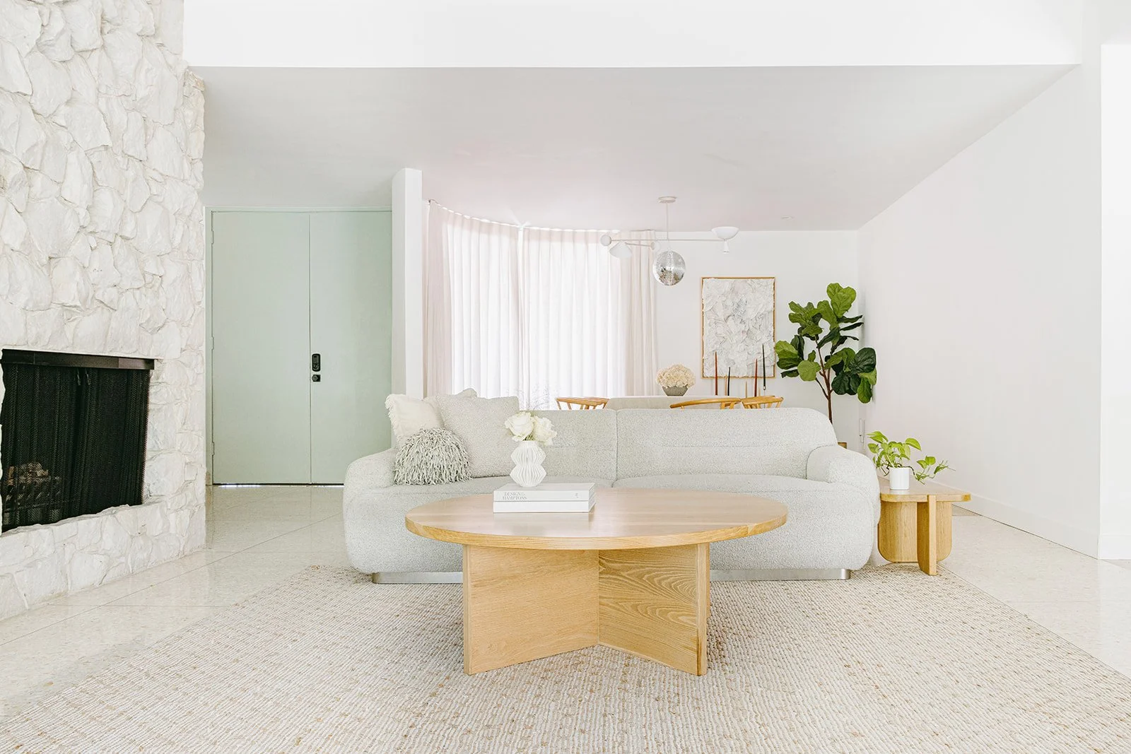 Bright living room with white walls, a light gray sofa, wooden coffee table, green plants, and a fireplace, with natural light coming through sheer curtains.