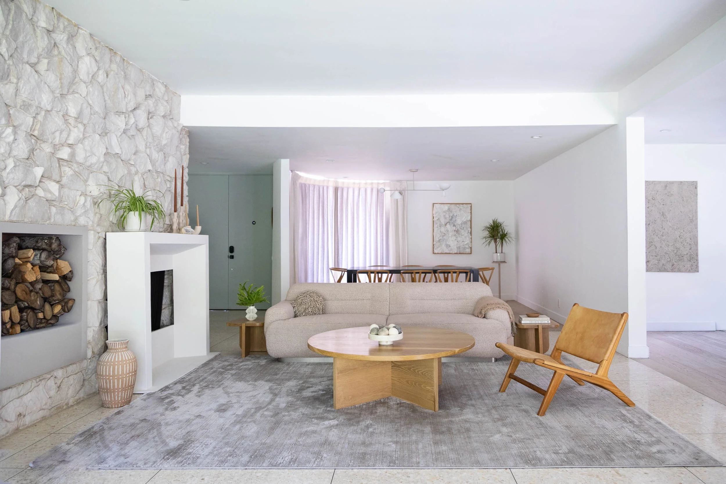 Modern living room with a beige sofa, a round wooden coffee table, a tan lounge chair, and a stone fireplace with firewood. The space is decorated with plants, art on the walls, and sheer curtains over large windows.
