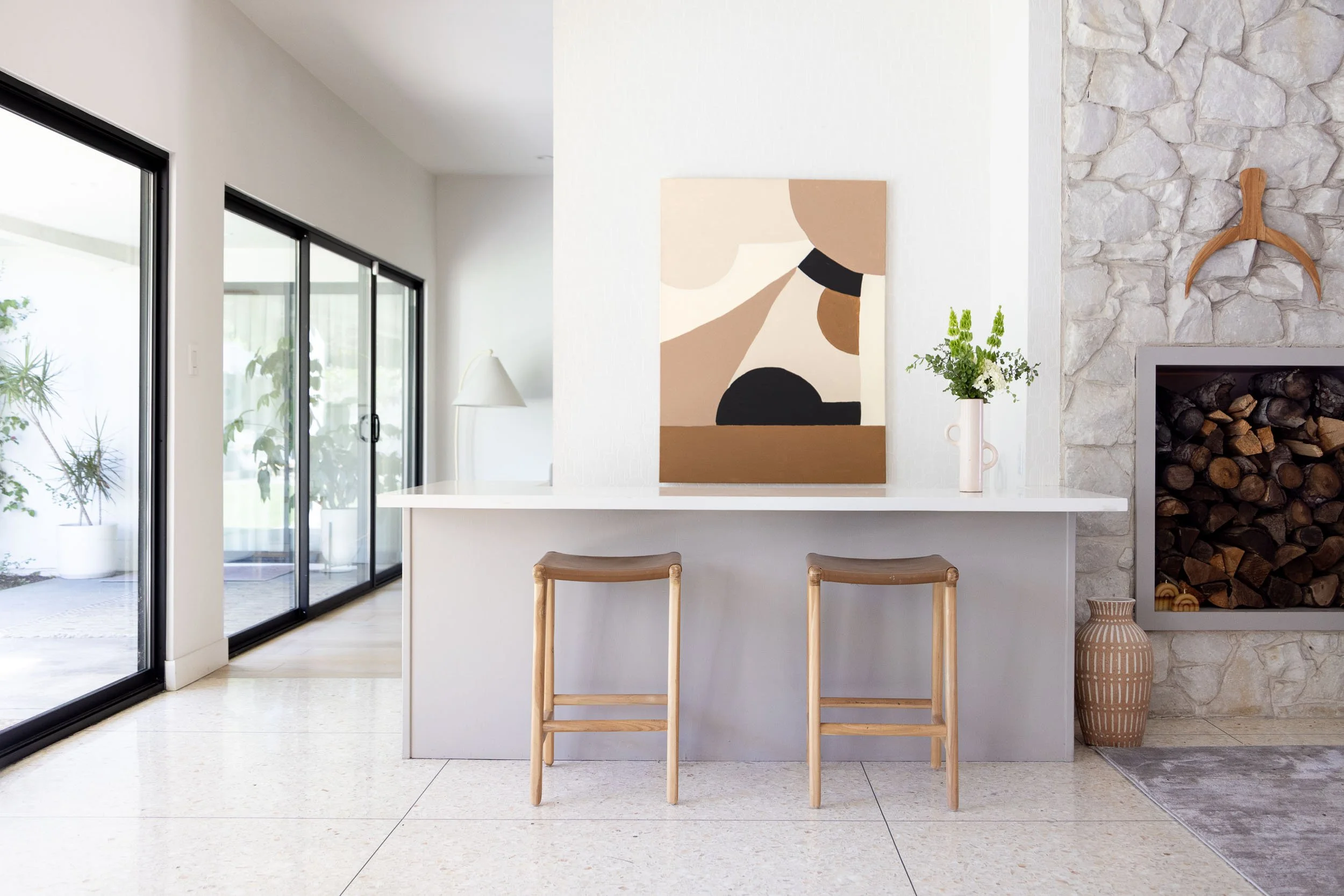 Minimalist modern dining area with a white bar counter, two wooden stools, and a piece of abstract art on the wall. There's a stone fireplace with a wood storage and decorative vase on the floor, and a sliding glass door leading outside with potted p