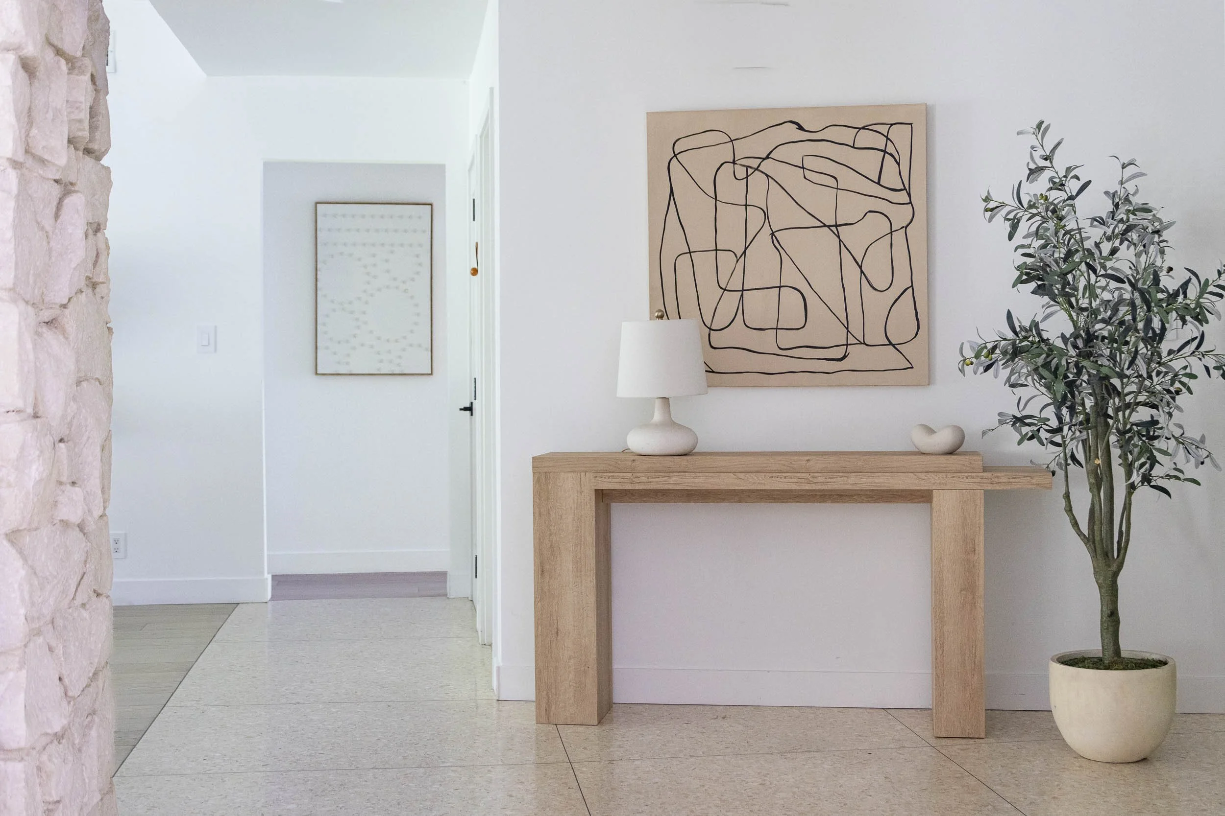 Minimalist interior with a wooden console table, white lamp, abstract black-line art painting, small sculpture, a potted tree, white walls, and minimalist decor.