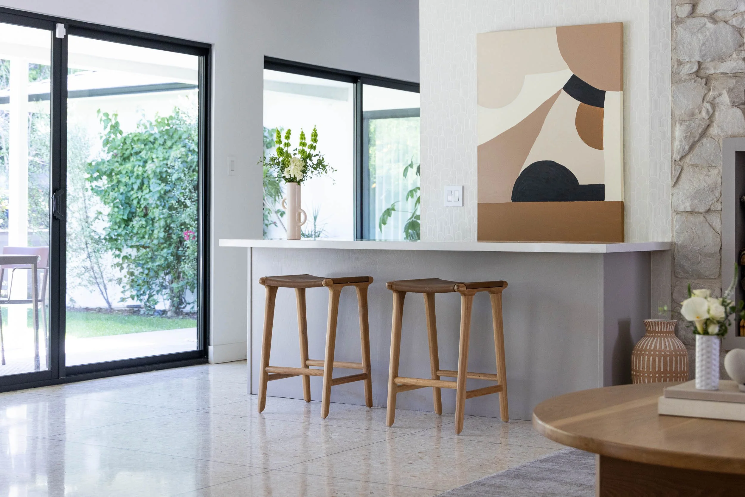 Modern living space with a white bar counter, two wooden stools, a large abstract painting with earth tones on the wall, a vase with flowers, and sliding glass doors leading outside with greenery visible.