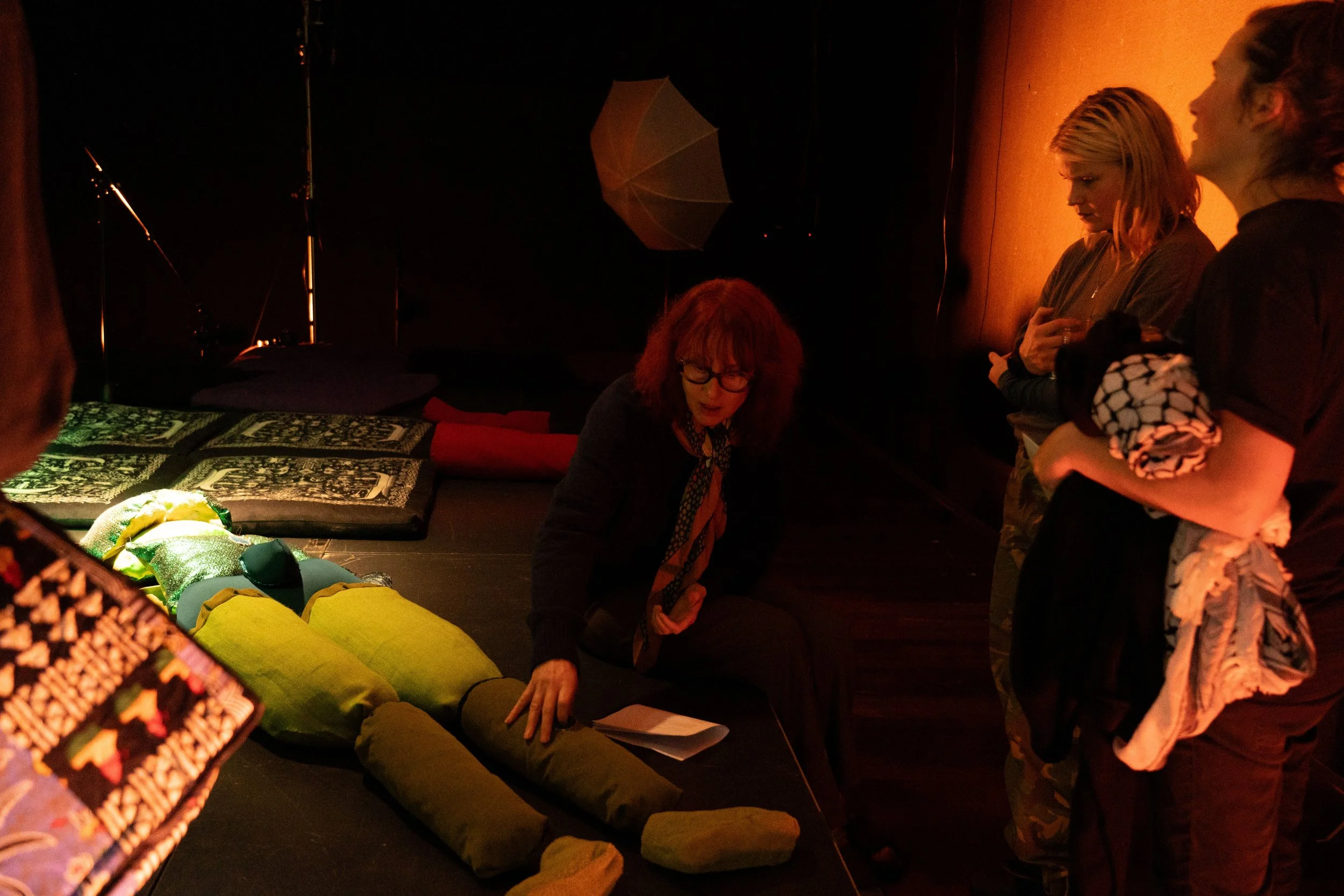Three people in a dark roon, looking at an art installation that’s on the ground. Workshop facilitator Sandra Lange is sitting next to it, and telling the others about the art. One person has an keffiyeh in their hand.