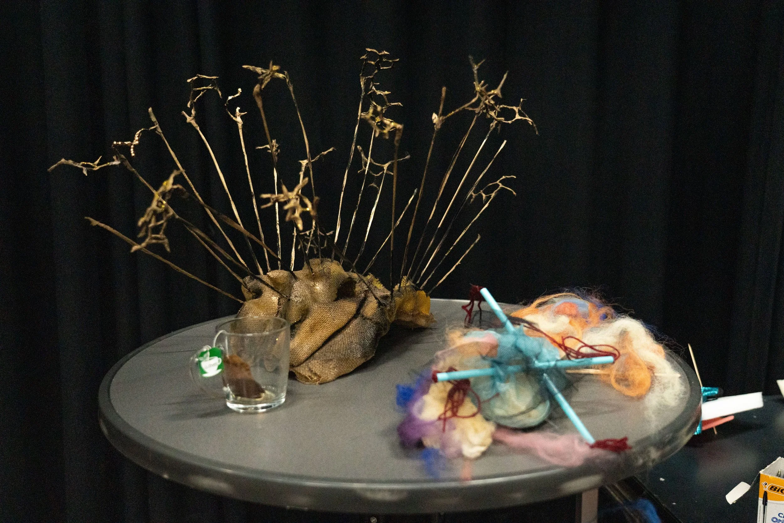 Pieces of art on a small round table, the art relates to pain. One pieve of art hase sharp spikes, the other has sticks with loose wool. Ther's an empty teacup.