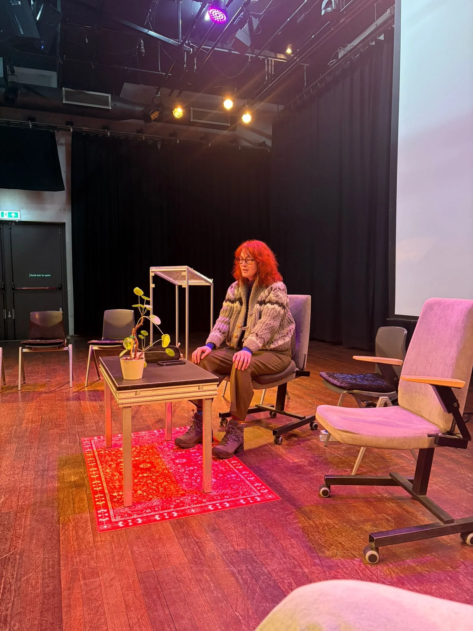 A androgynous person with red frizzy hair, a cozy hiking outfit and cat-eyed glasses sits on a chair. Alone, in a circle of empty chairs. They look tired, with their hands on their knees.