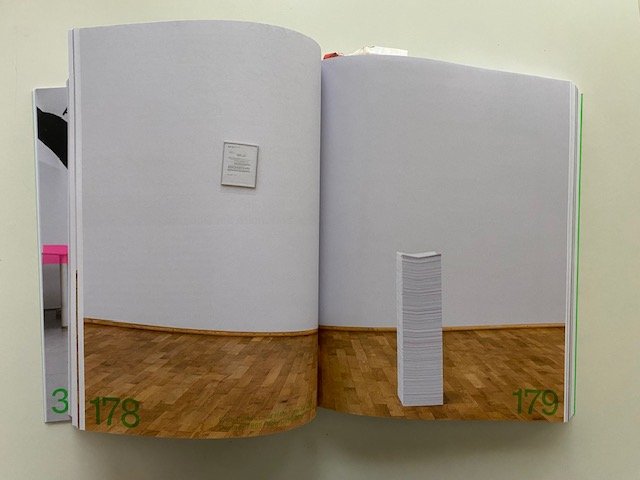 An exhibition space at MMK, wood flooring, white walls. On the wall hangs a framed hospital invoice and in the middle of the space there is a stacked pile of 7865 white papers; medical records, bills, and treatment plans.