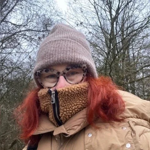 Woman standing in forrest on cold winter morning. Face barely visible behind big glasses, eyes look happy. Long red hair, wool cap and thick winter coat, and fleece sweater.