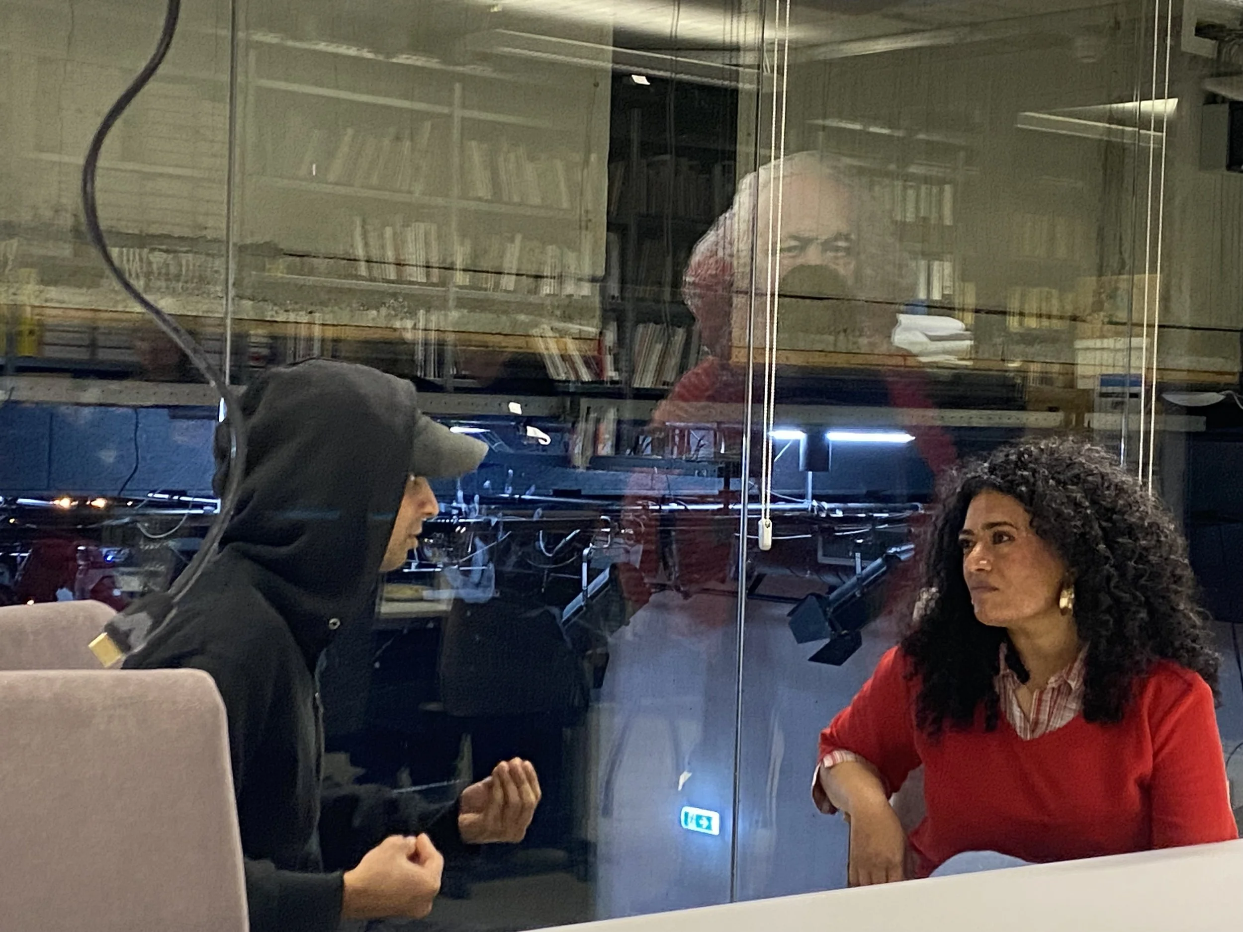 2 persons talking to each other. They're sitting before a glass wall. In the reflections of the glass there's a big silhouette of Karl Marx <3