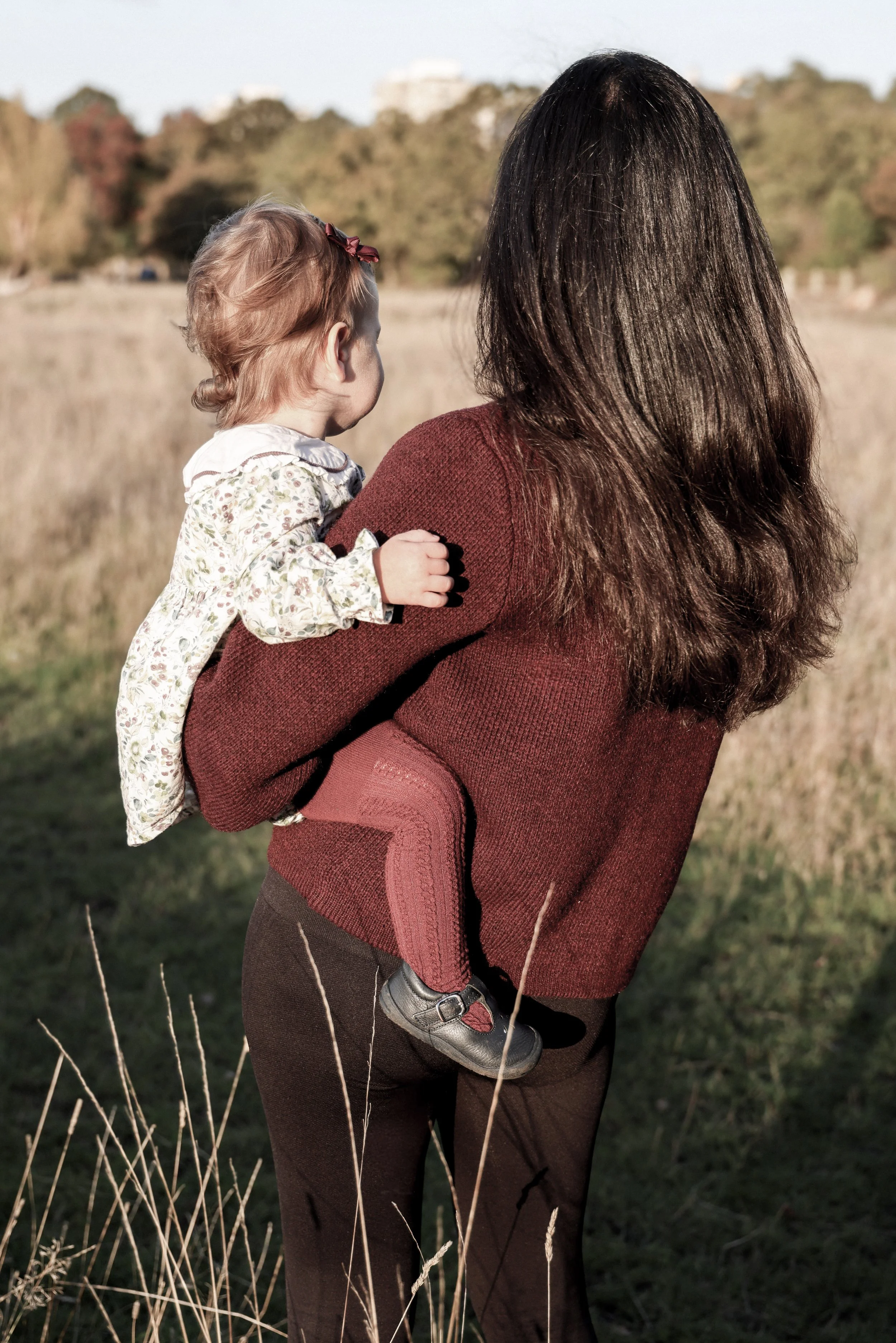 A woman with long brown hair holding a young girl with light curly hair, both standing outdoors in a grassy field with trees in the background, during the daytime.