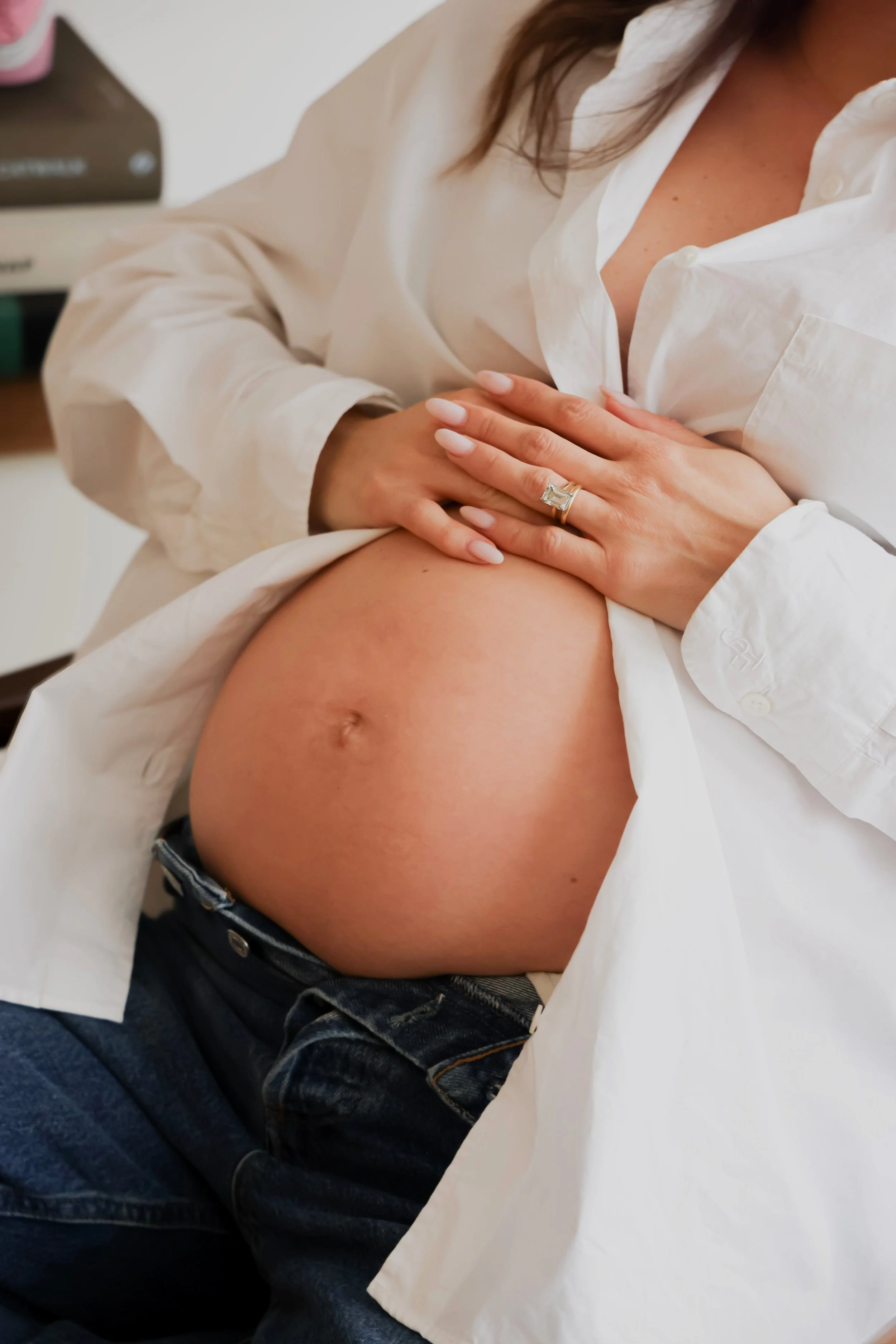 Close-up of a pregnant woman's belly with her hands resting on her chest, wearing a white shirt and a wedding ring.