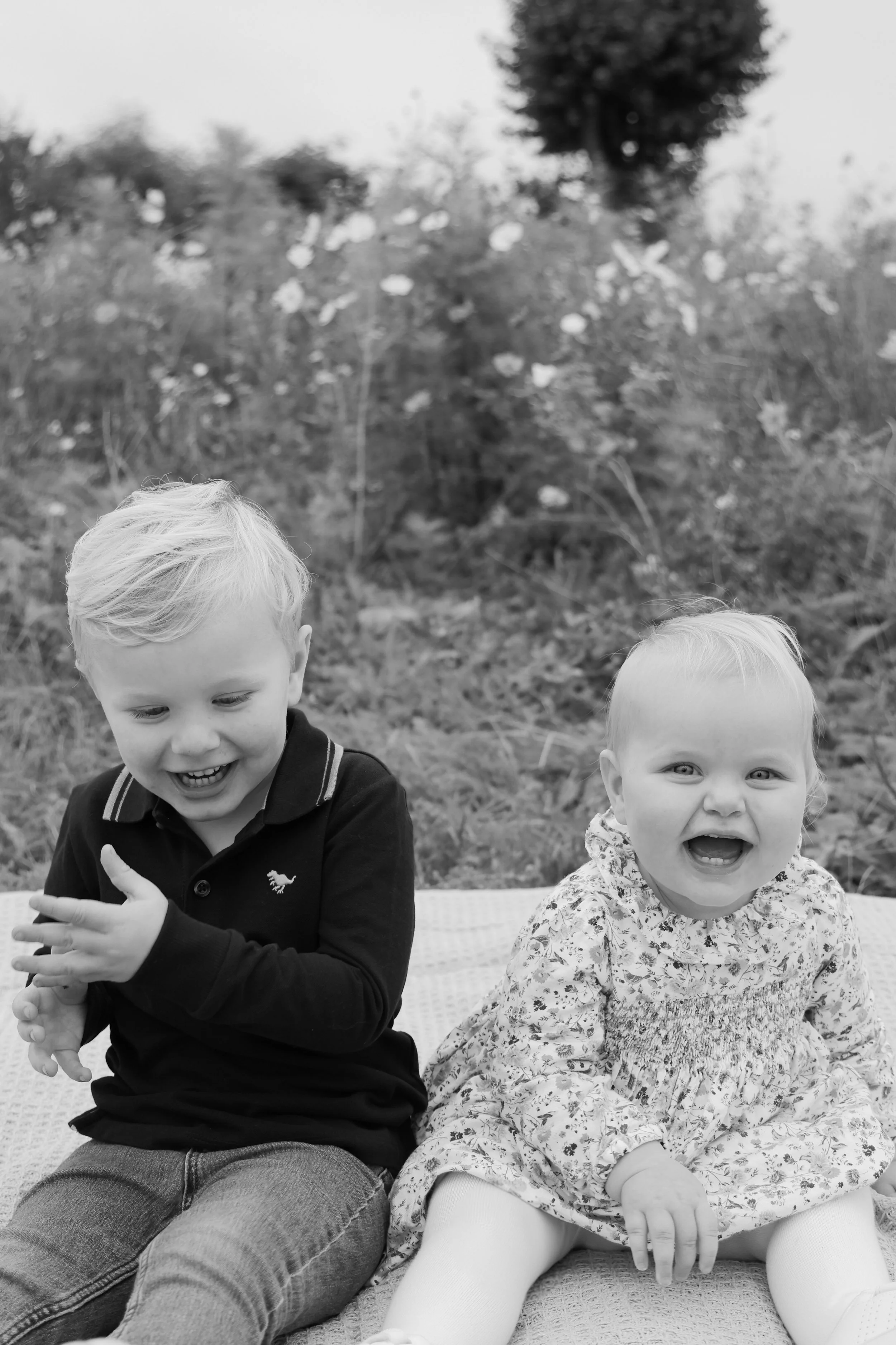 Two young children, a boy and a girl, sitting outside on a blanket, smiling and laughing with a garden and trees in the background, in black and white.
