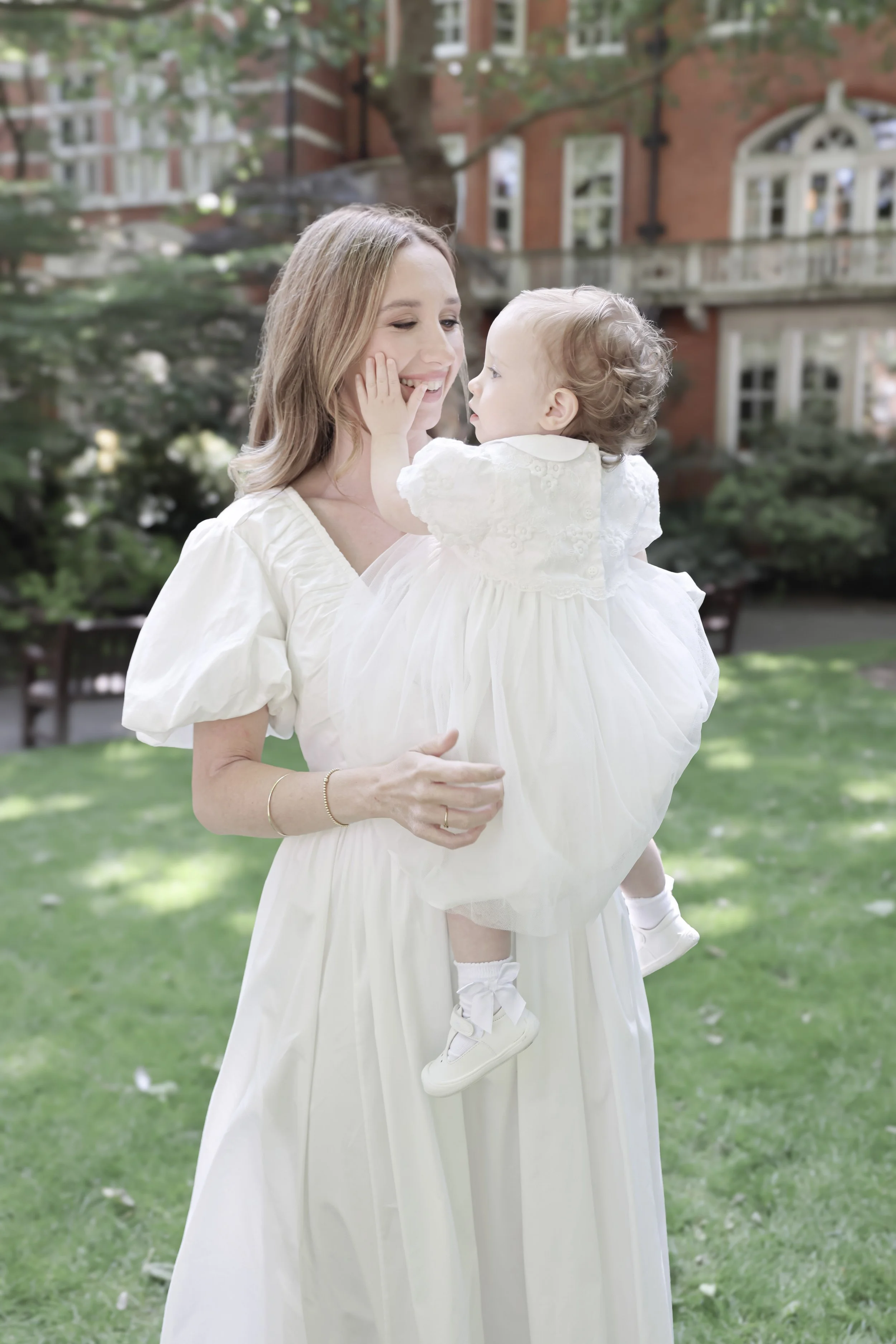 A woman in a white dress holding a young girl in a white dress and sneakers, both smiling in a park area with grass and trees.