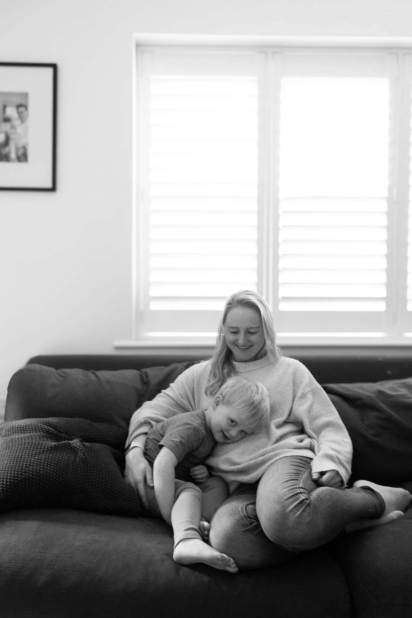 A black and white photo of a woman and a young boy sitting on a sofa in a living room. The woman is smiling and the boy is playfully leaning on her, looking at the camera. There is a window with blinds behind them and a framed picture on the wall.