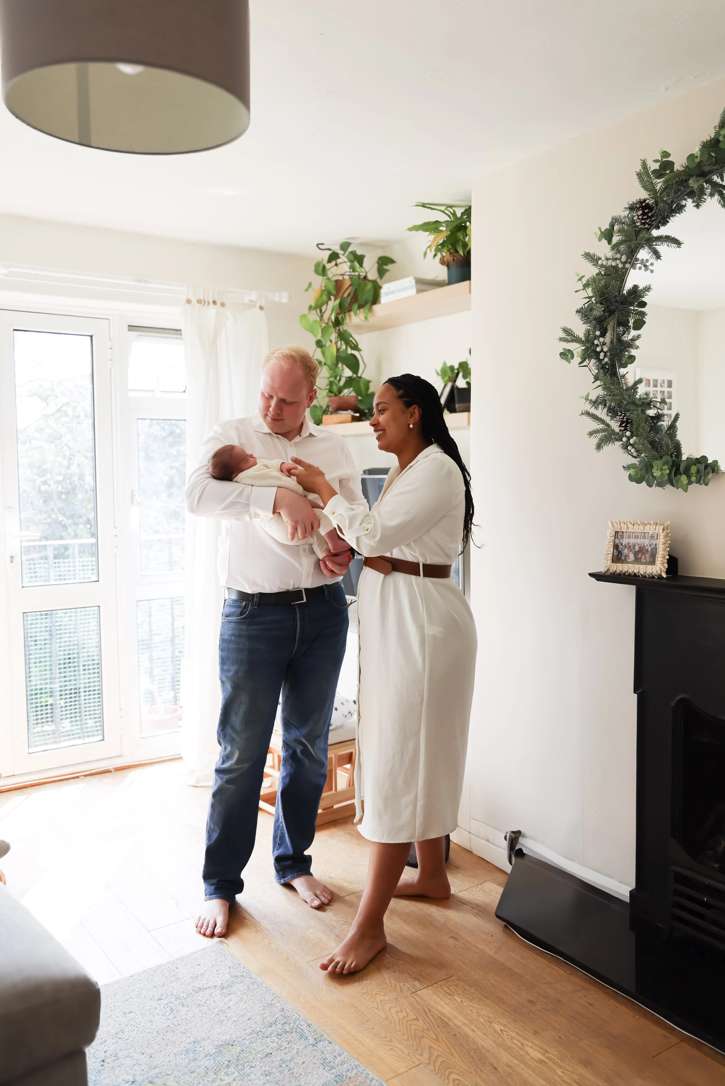 A couple holding a newborn baby near a fireplace in a bright living room decorated with plants and a framed photo.