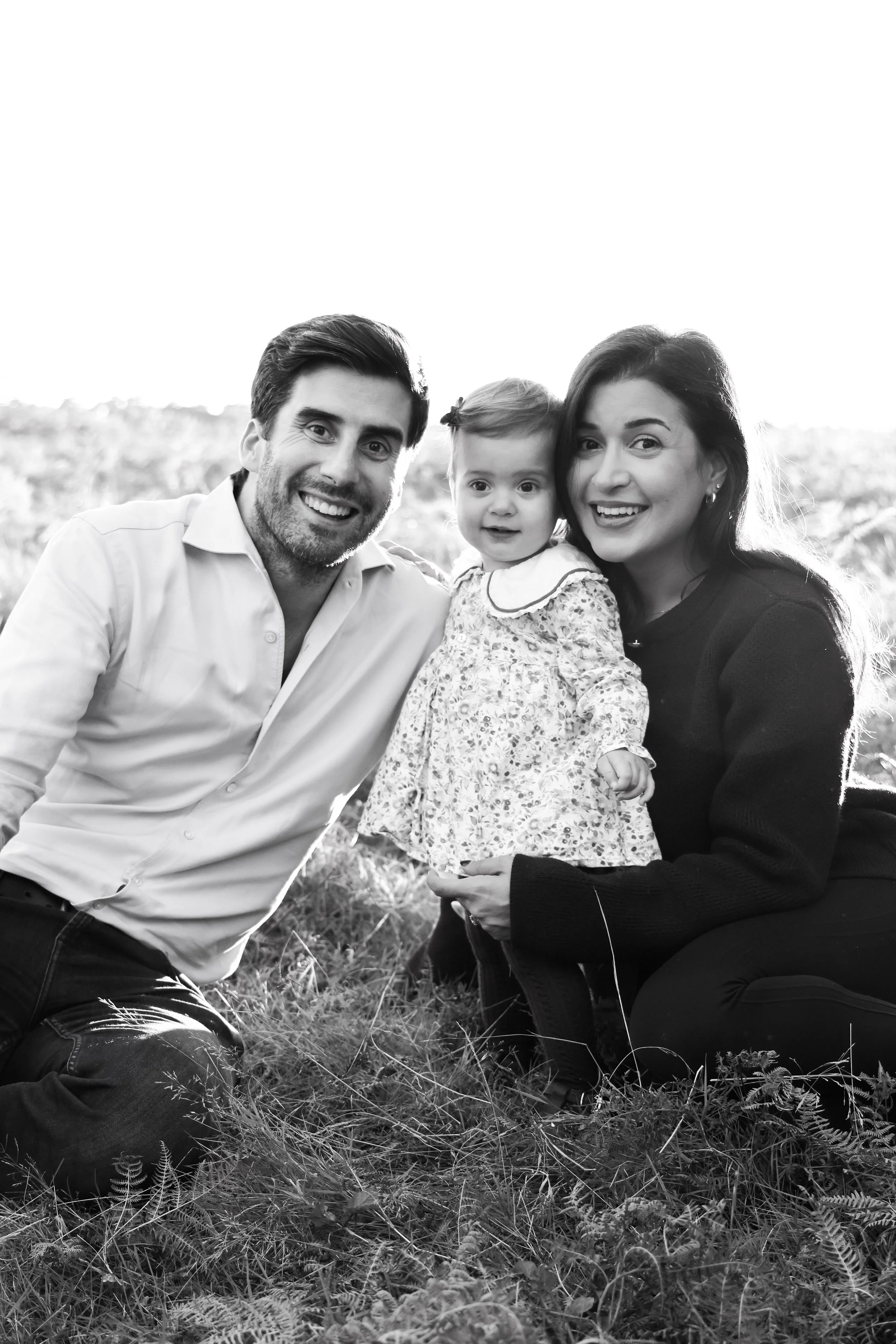 A black and white photo of a family of three outdoors, with a man, woman, and a young girl. They are smiling and close together, sitting on grass.