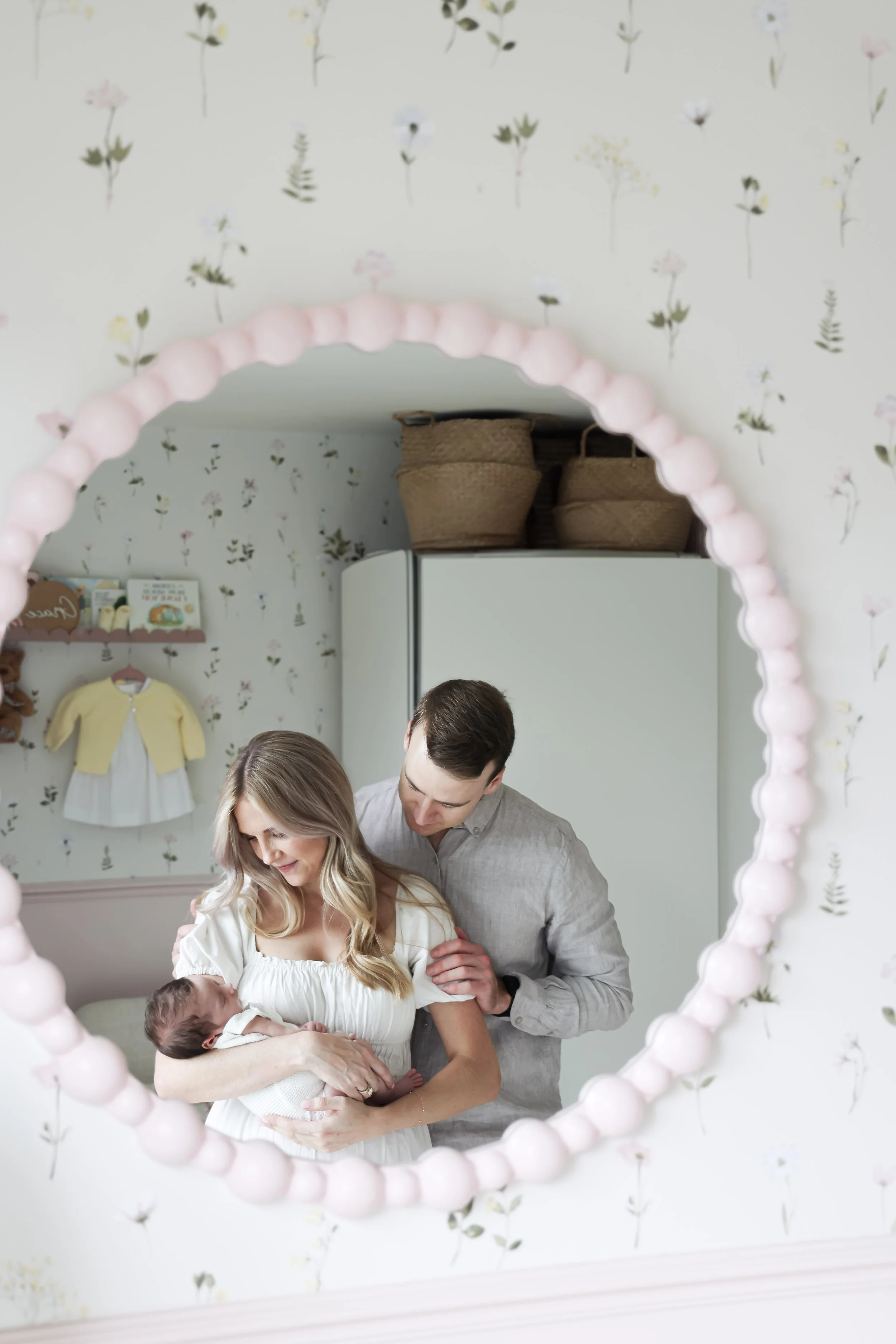 A reflection in a round pink beaded mirror shows a woman holding a newborn baby, with a man standing behind her, in a room with floral wallpaper and children's clothing on the wall.