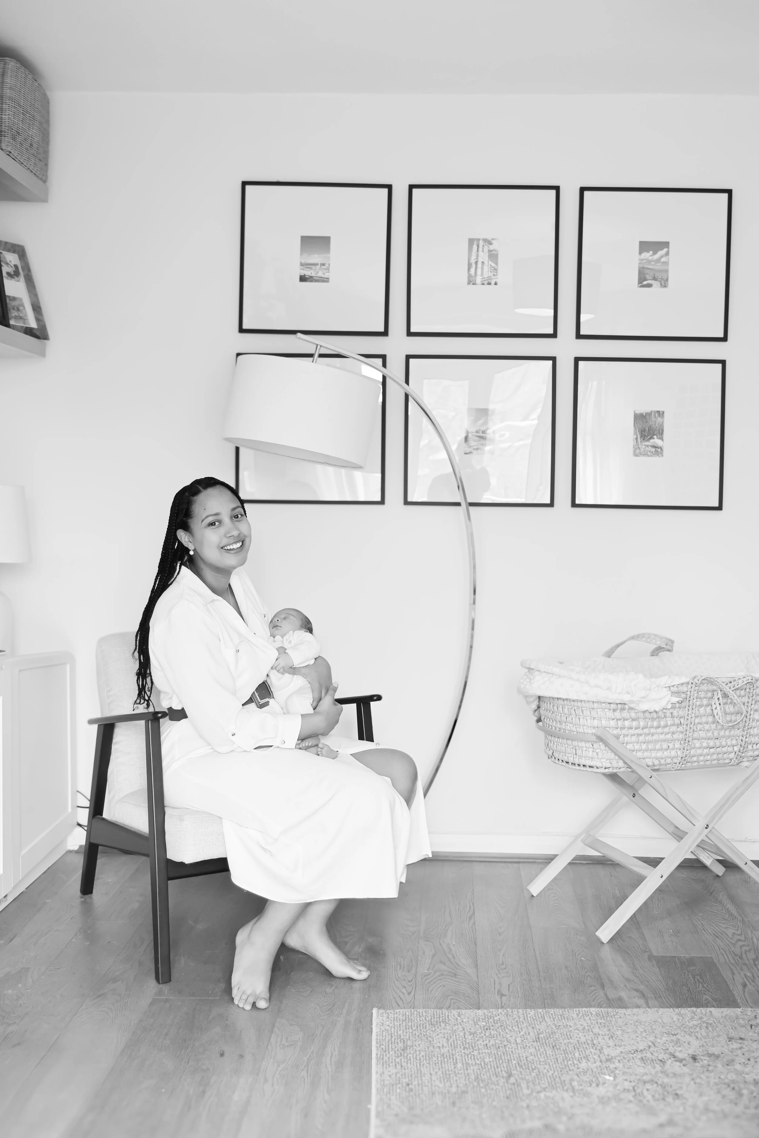 A woman sitting on a chair, holding a newborn baby, smiling at the camera in a modern, well-decorated room with framed pictures on the wall and a crib nearby.
