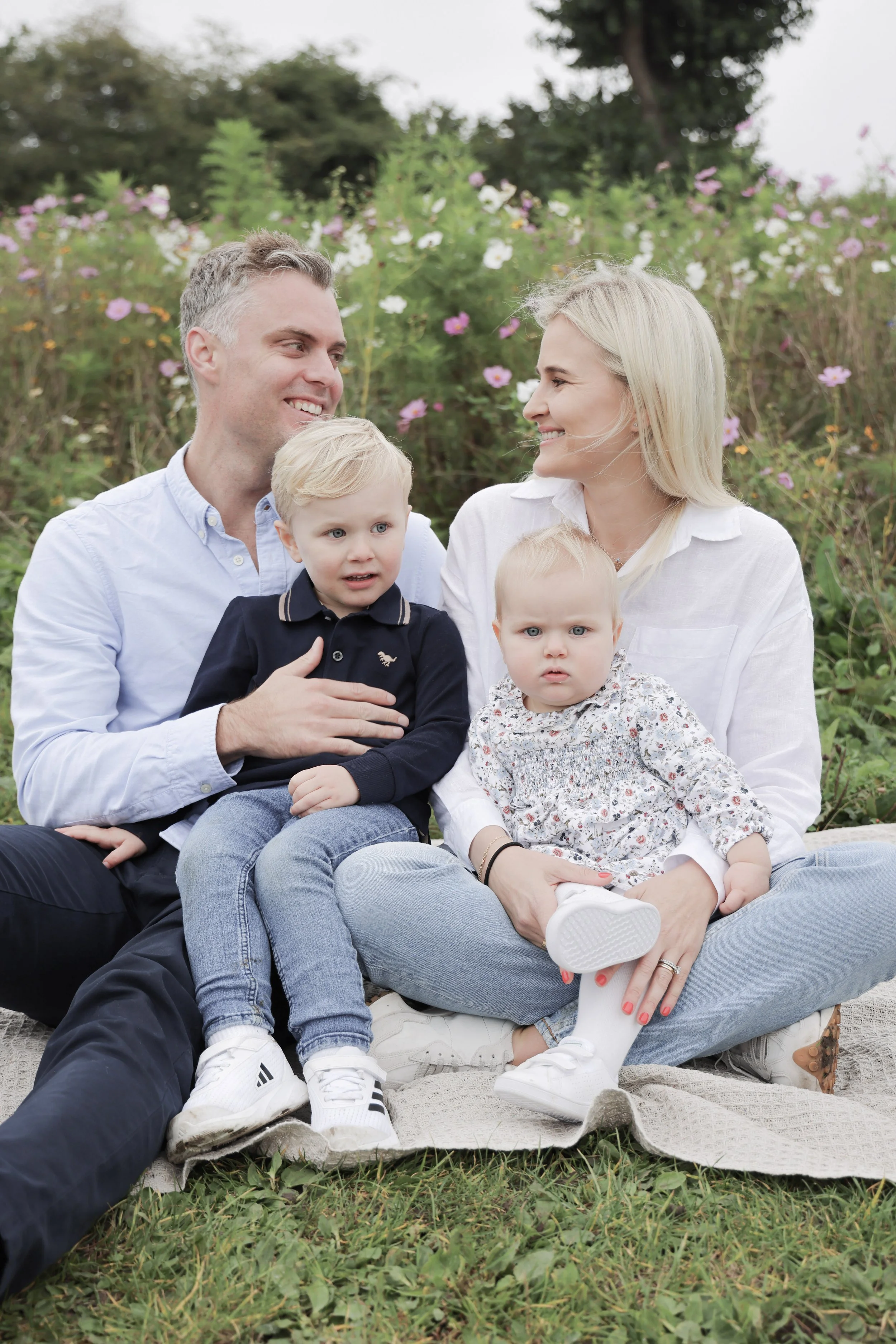 A happy family of four sitting on a blanket outdoors in a garden with colorful flowers. The father and mother are smiling at each other. The young boy is sitting on his father's lap, looking slightly to the side. The little girl is sitting on her mot
