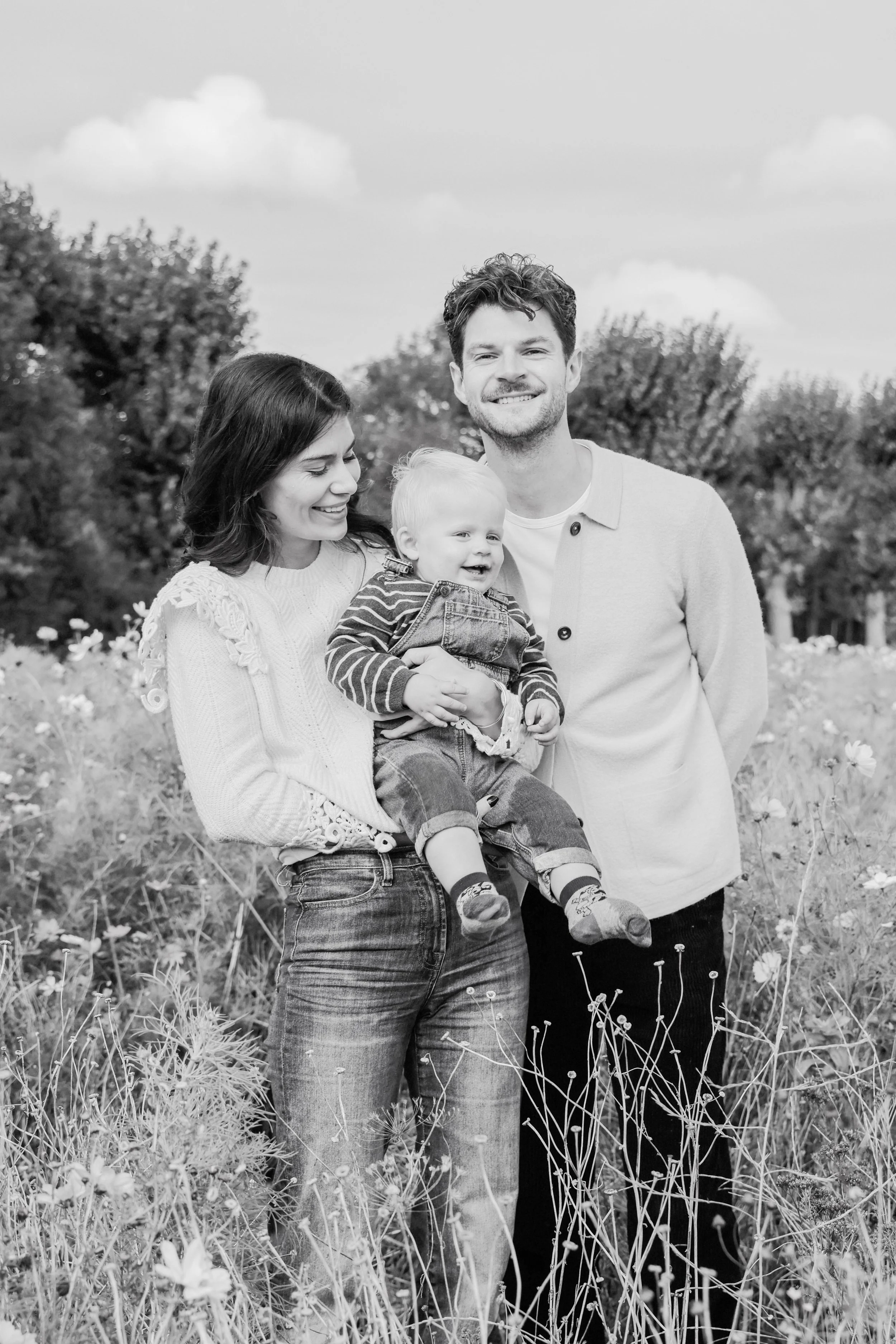 A black-and-white photo of a smiling family of three standing outdoors in a field of wildflowers with trees and clouds in the background. The mother has dark hair and is holding a small child, while the father stands beside them.