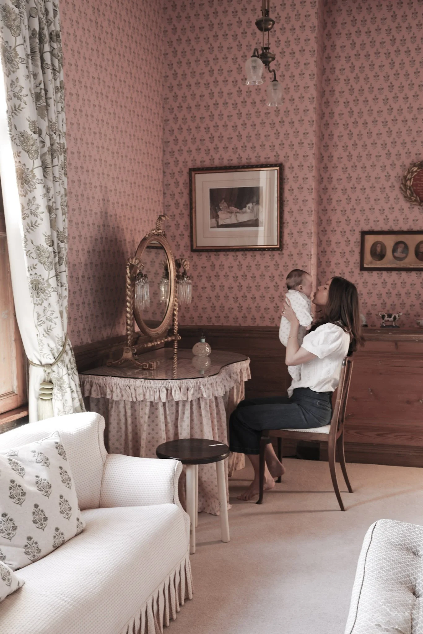 A woman sitting on a wooden chair holding a baby girl, standing in a vintage-style room with pink floral wallpaper, a mirror, framed pictures, and a chandelier.