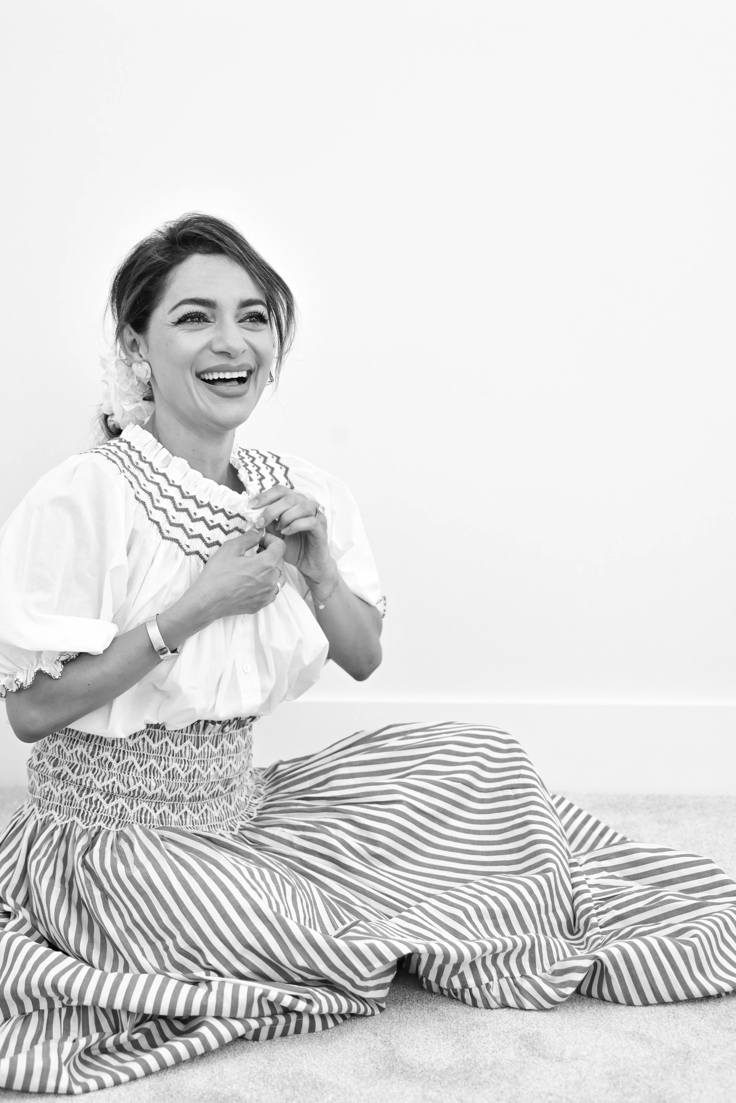 A smiling woman sitting on the floor, wearing a white blouse and a striped skirt, holding her hands near her chest.