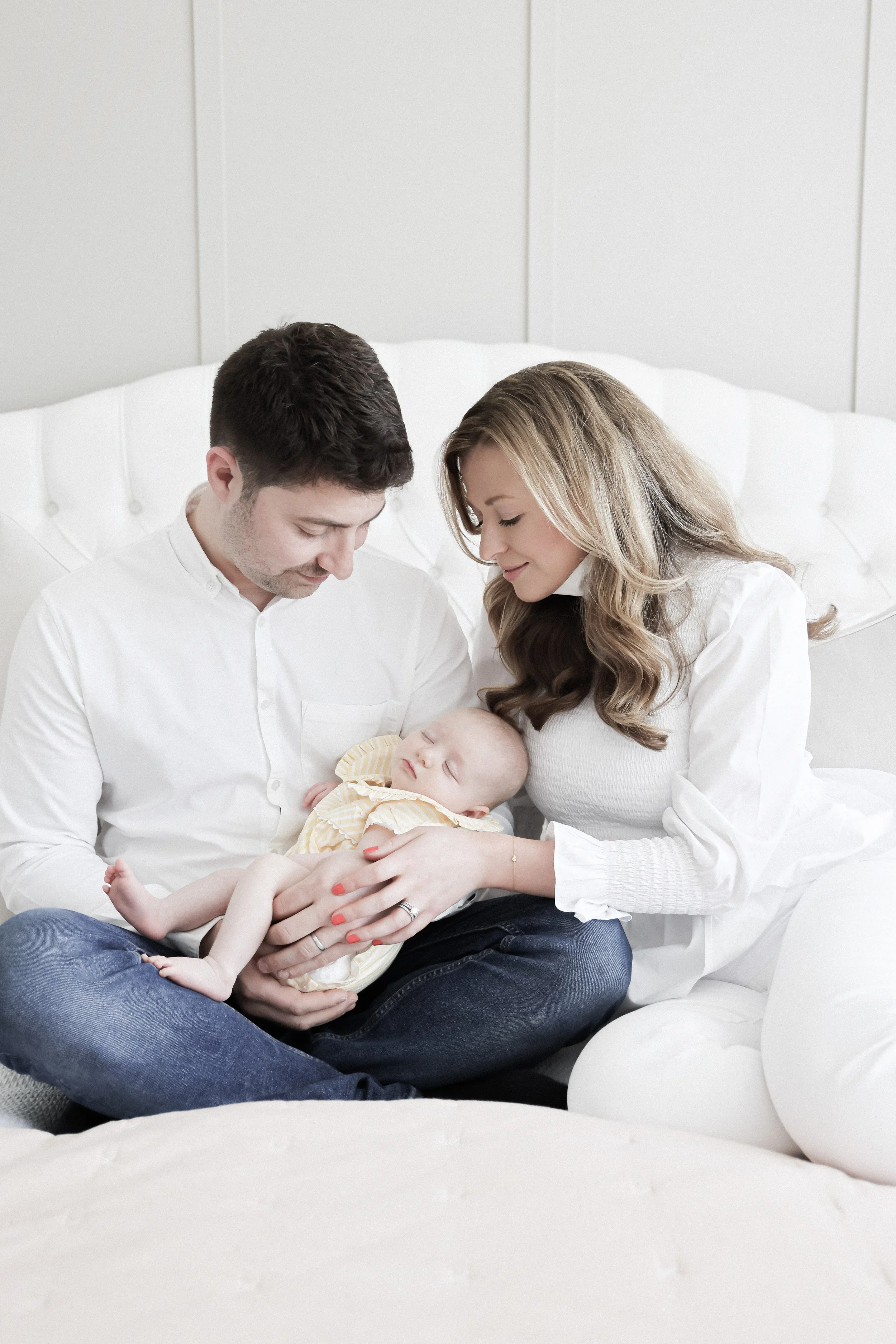 A young family sitting on a bed with a white headboard, holding a sleeping baby, all smiling softly at the baby.