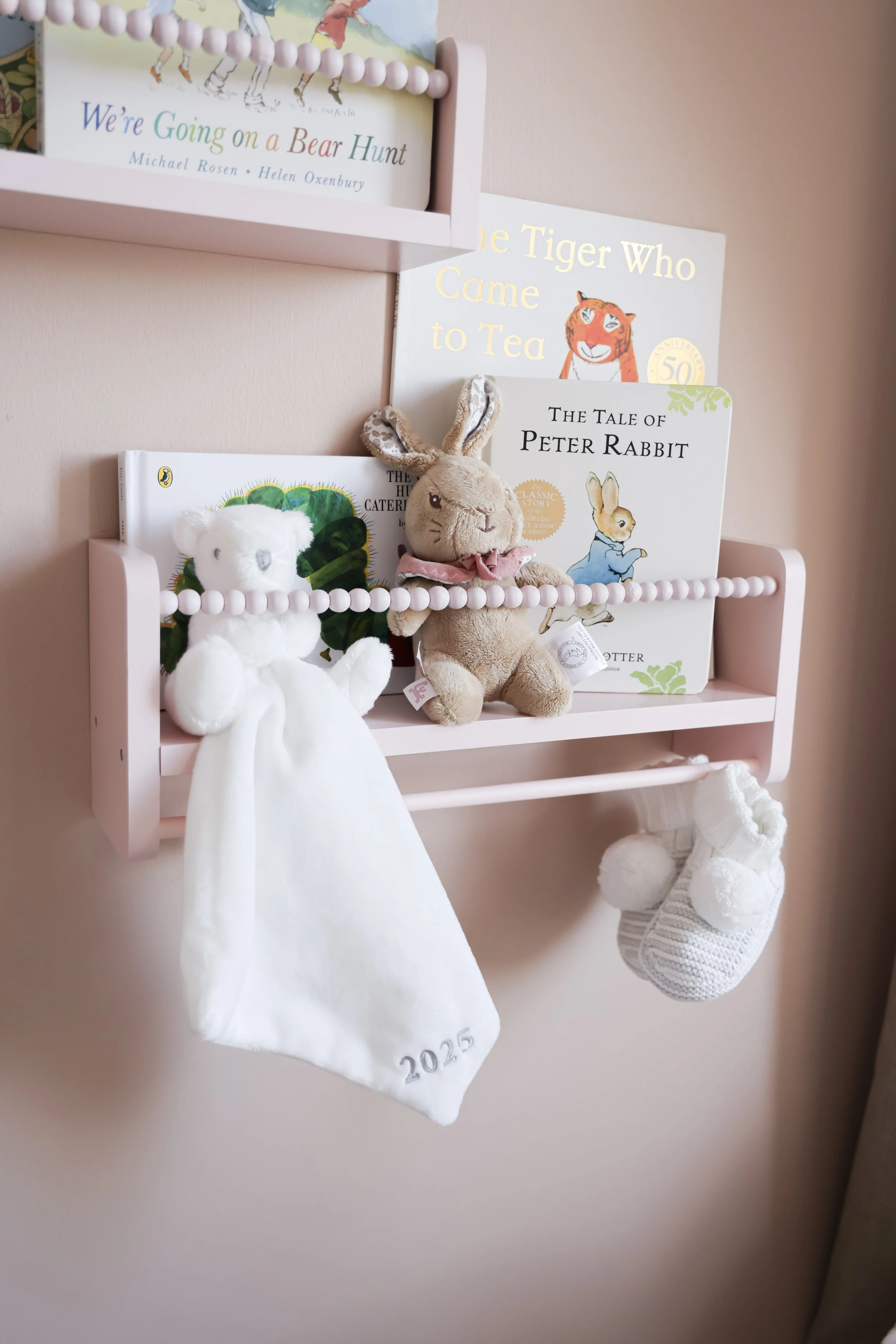 Pink wall-mounted shelf holding children's books and plush toys, including a bunny and a white teddy bear, with a hanging sock reading '2025' and a hanging white knitted stocking with pom-poms.