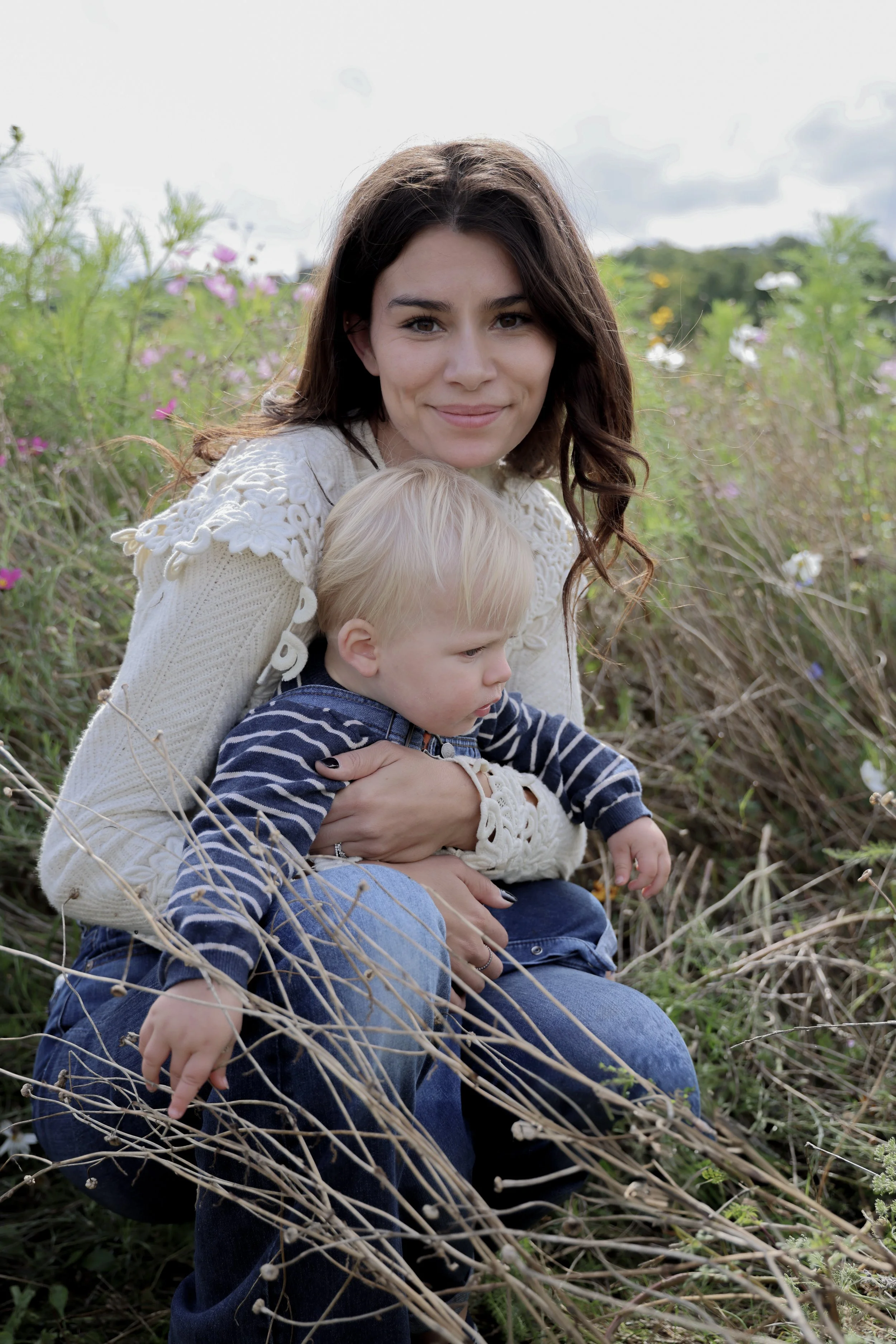 A woman with dark brown hair and a young blonde boy sitting outdoors in a field of wildflowers.