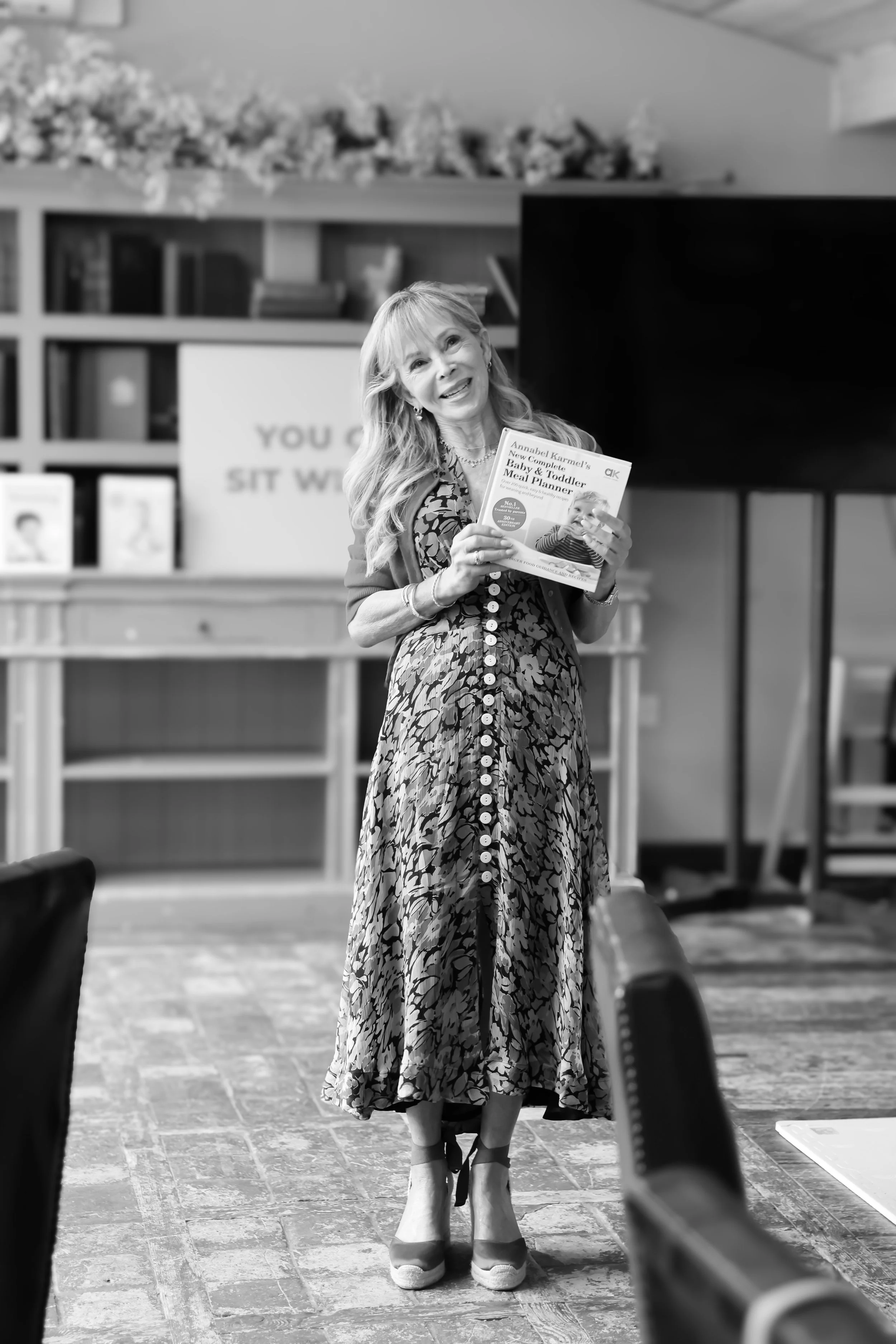 A smiling woman with long, wavy hair holding a book in a classroom or event space.