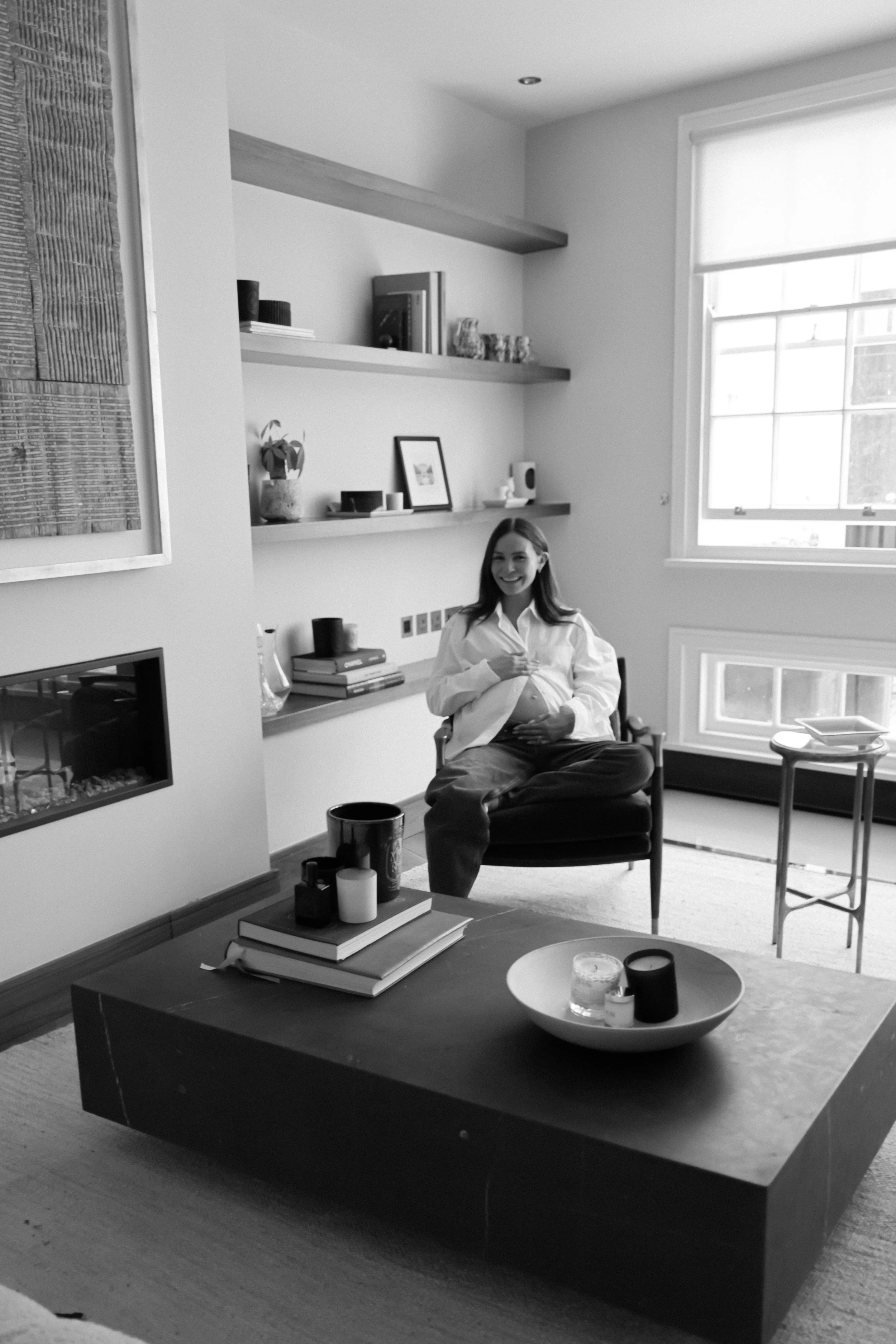 A woman with long hair sitting in a modern living room chair, smiling, with a large window behind her. A coffee table in front has candles and books, and shelves on the wall behind hold various decorative objects.