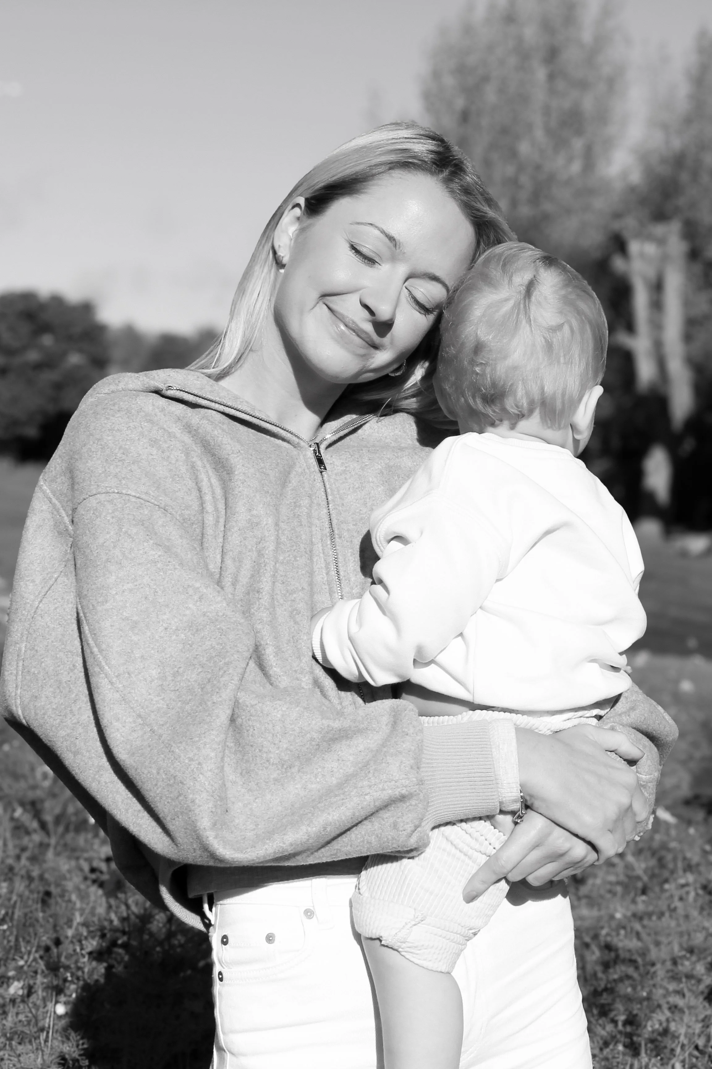 A woman with closed eyes and a smiling face holding a young boy outdoors in a park, with trees in the background.